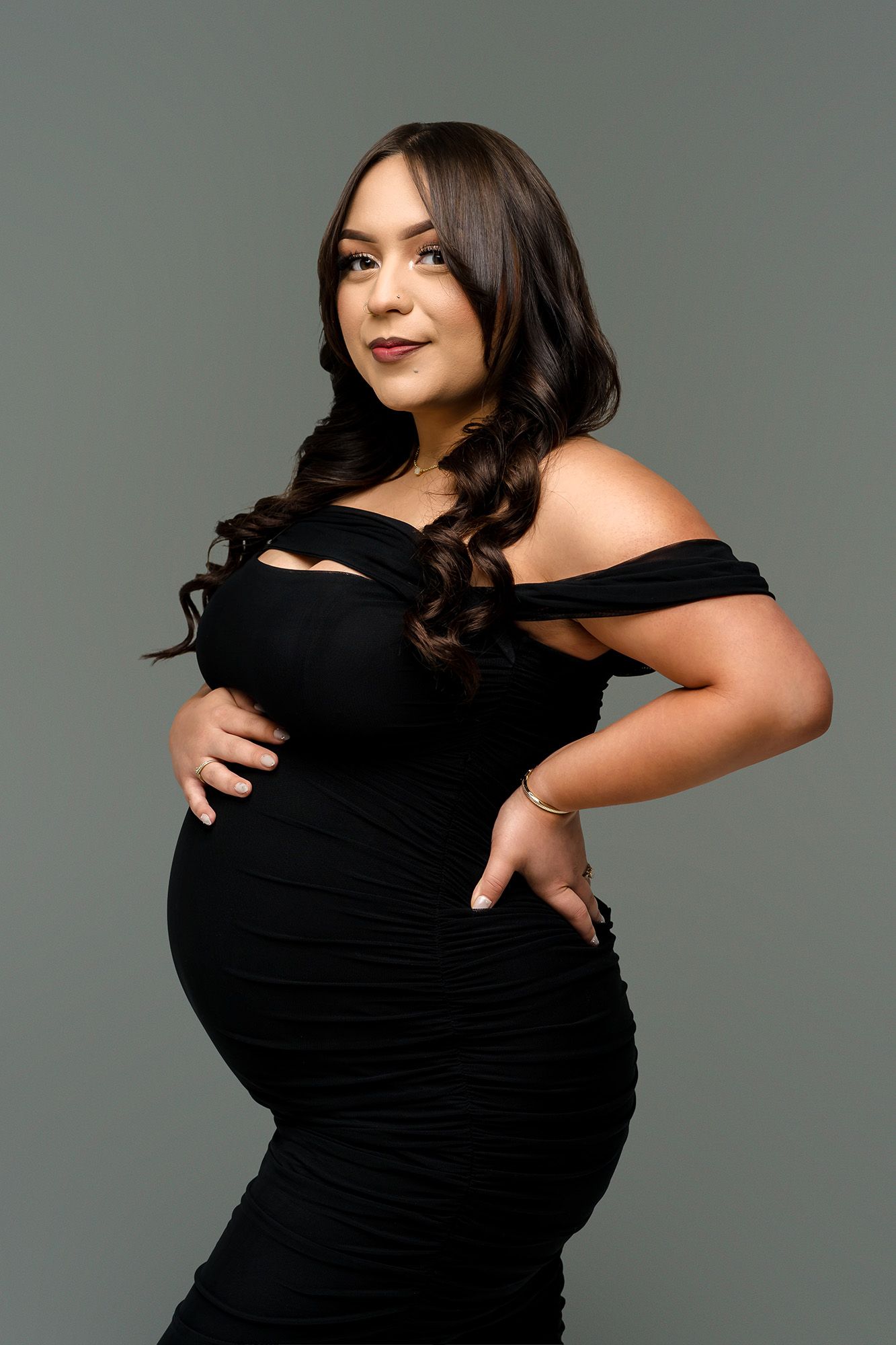 Pregnant woman in black off-shoulder ruched dress posing elegantly against gray background.