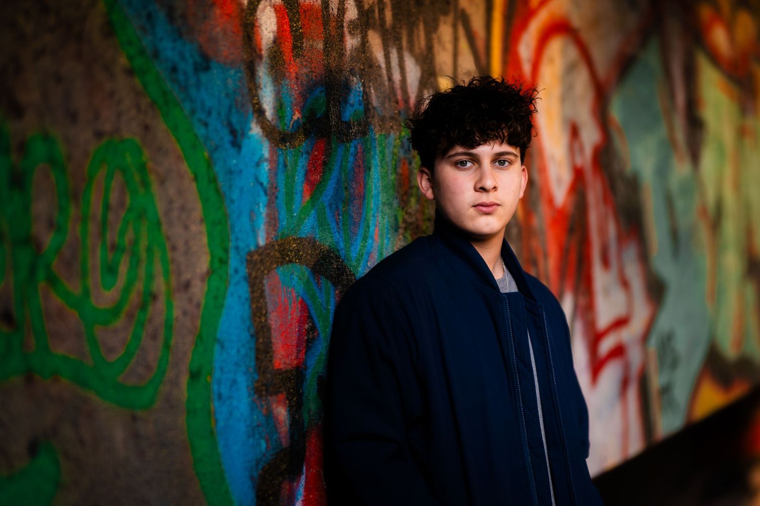 Person in dark jacket poses against colorful graffiti wall with dramatic lighting and vibrant urban backdrop.