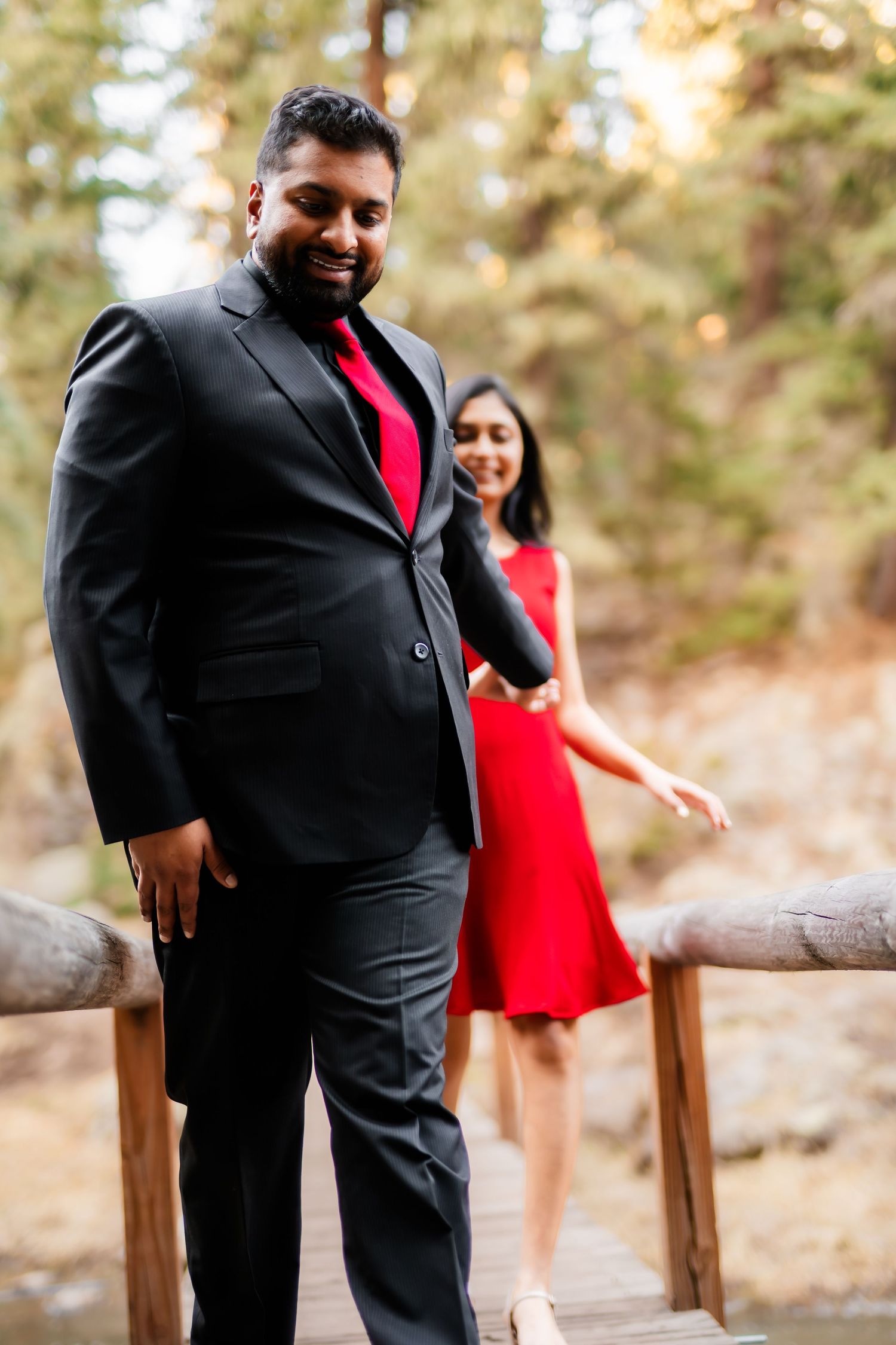 A couple in coordinating black and red formal wear poses on a wooden bridge.