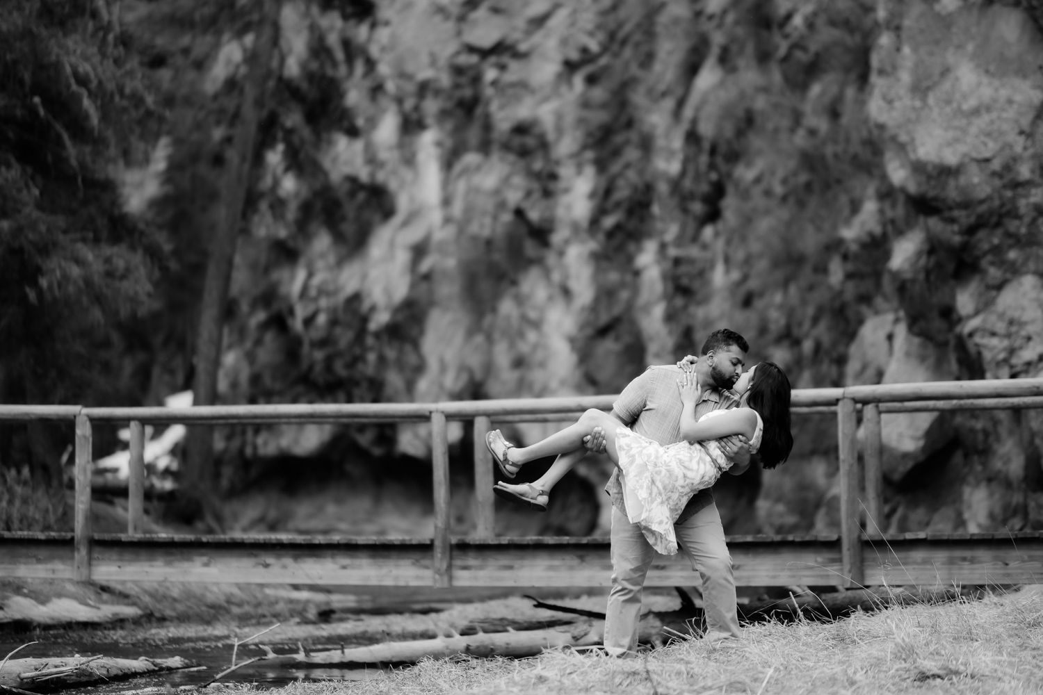 Couple shares romantic dance pose on wooden bridge with dramatic cliffside backdrop.
