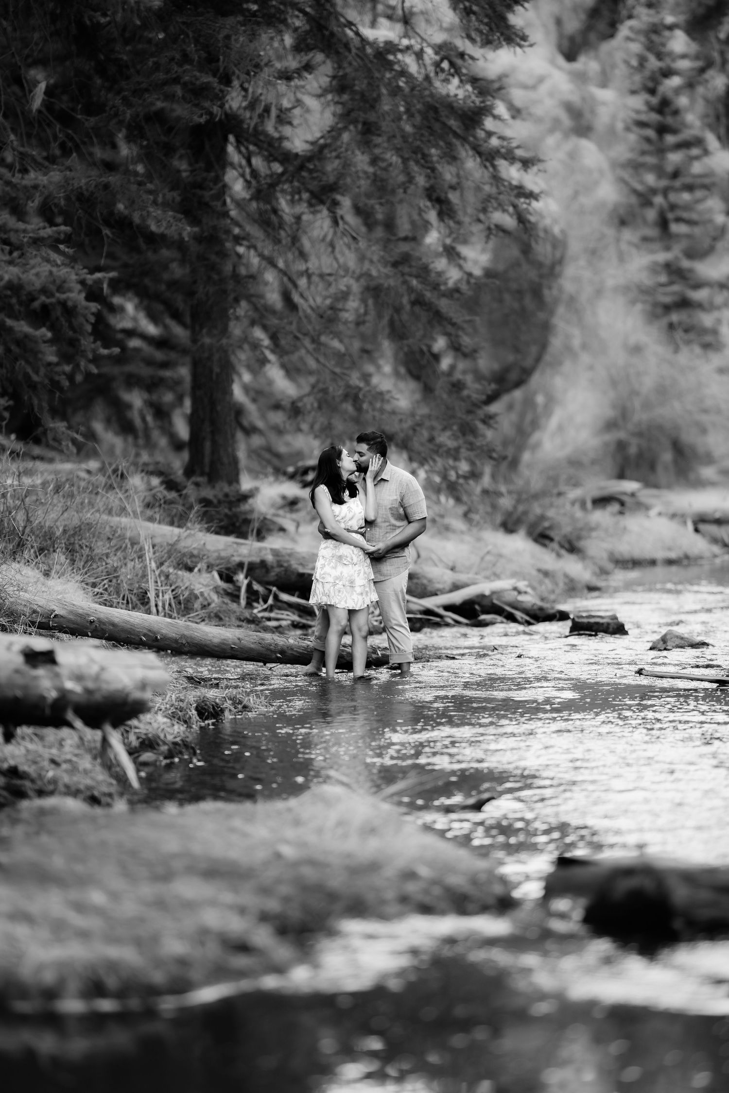 A couple shares a romantic moment by a stream in a misty forest setting.