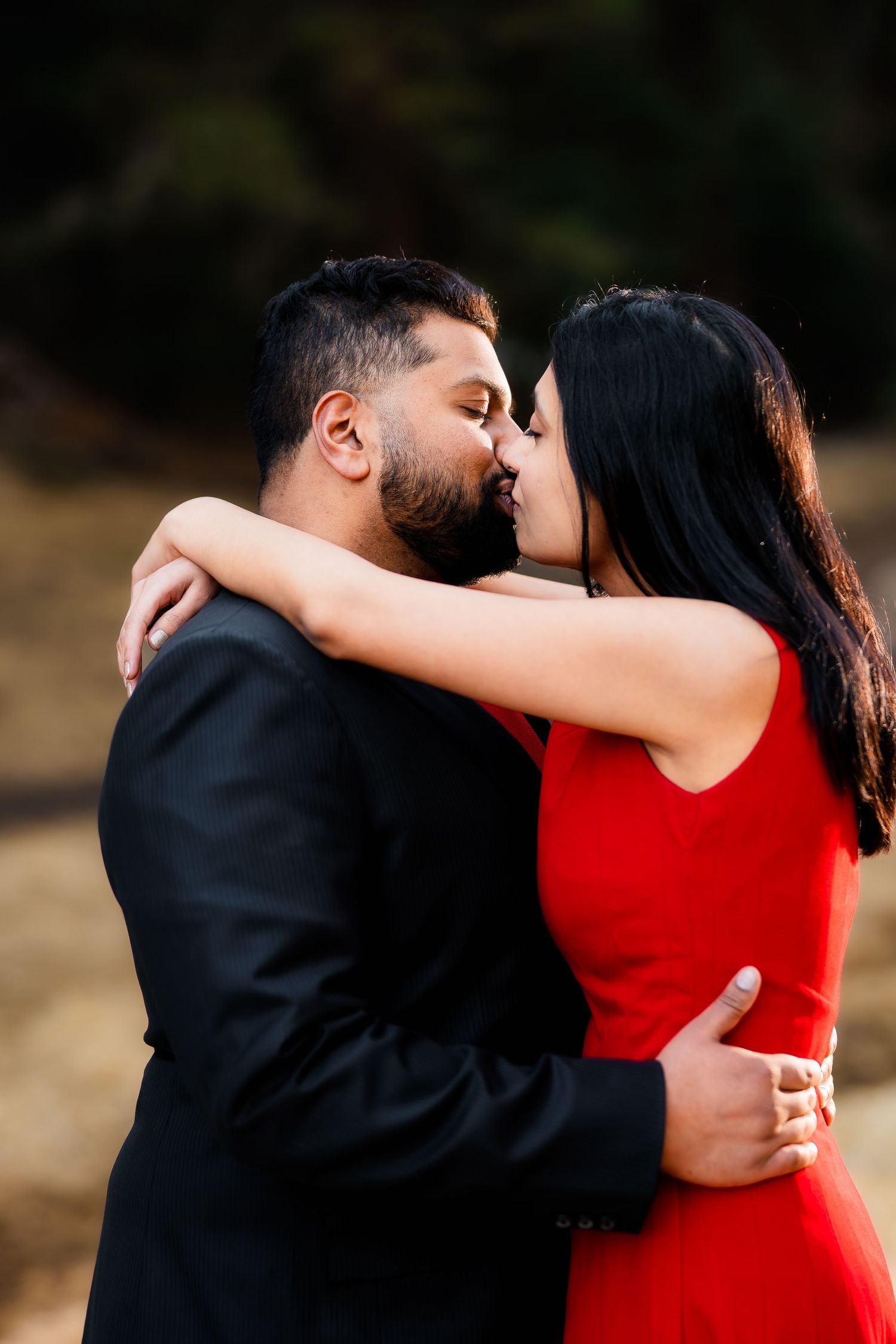 A romantic couple sharing a kiss, with the woman in a red dress and man in a black suit embracing outdoors.