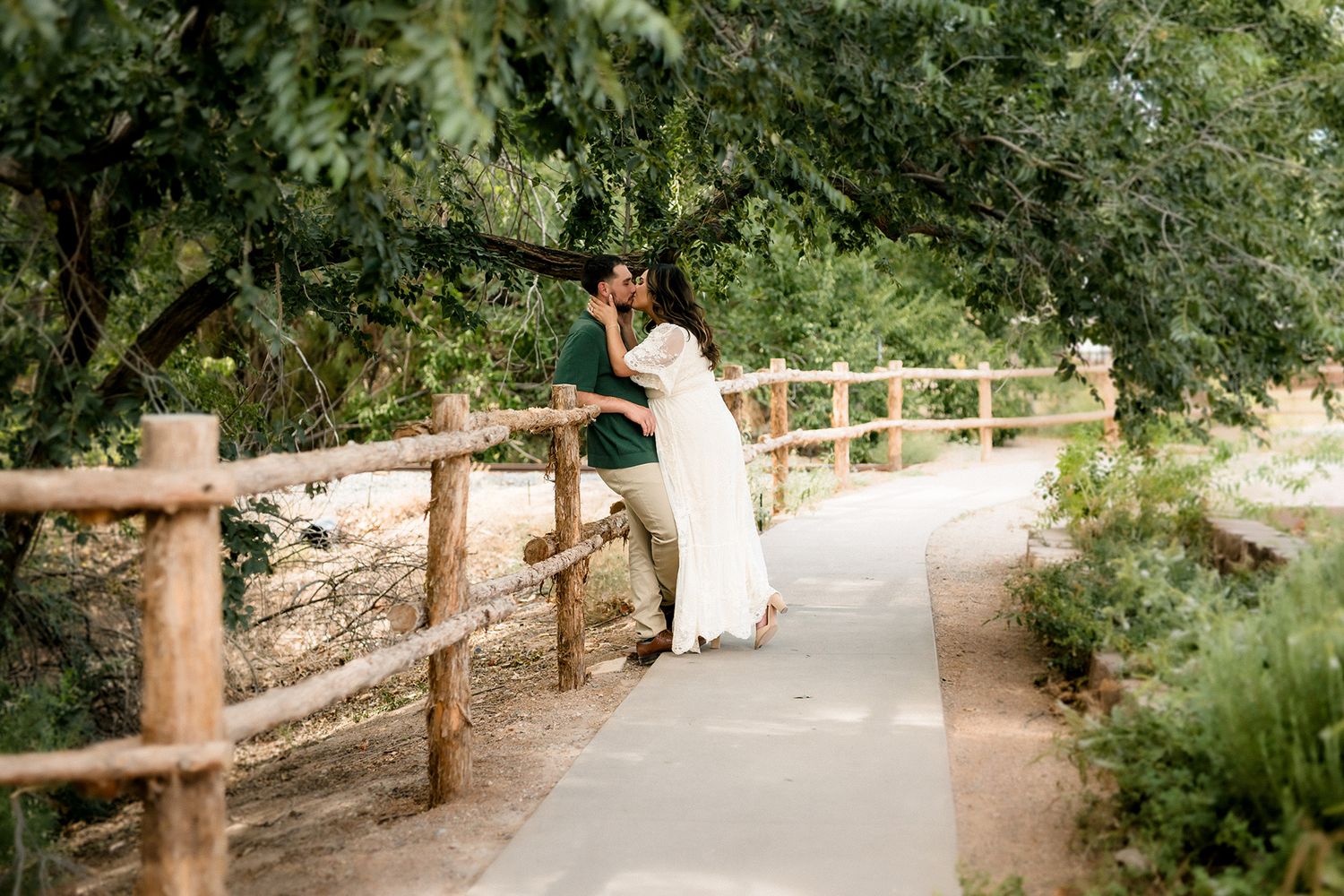 Couple sharing a romantic kiss on a rustic wooden bridge surrounded by lush green trees in an outdoor wedding setting.