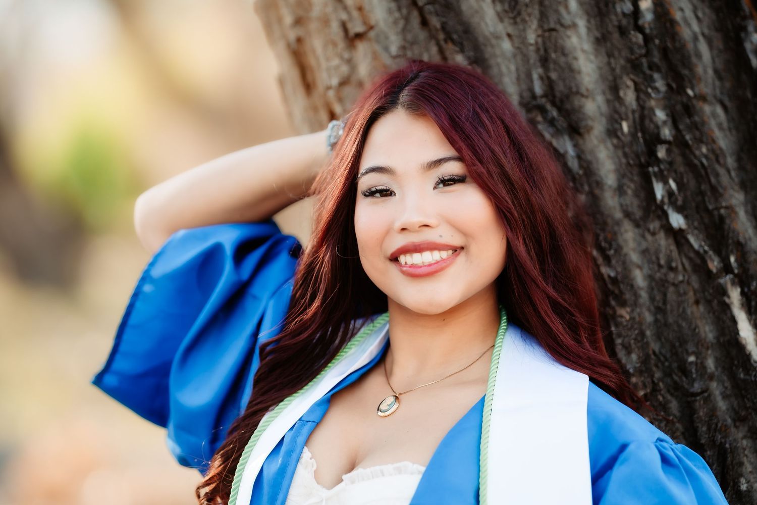 Graduate wearing blue and white graduation gown smiles while posing against tree.