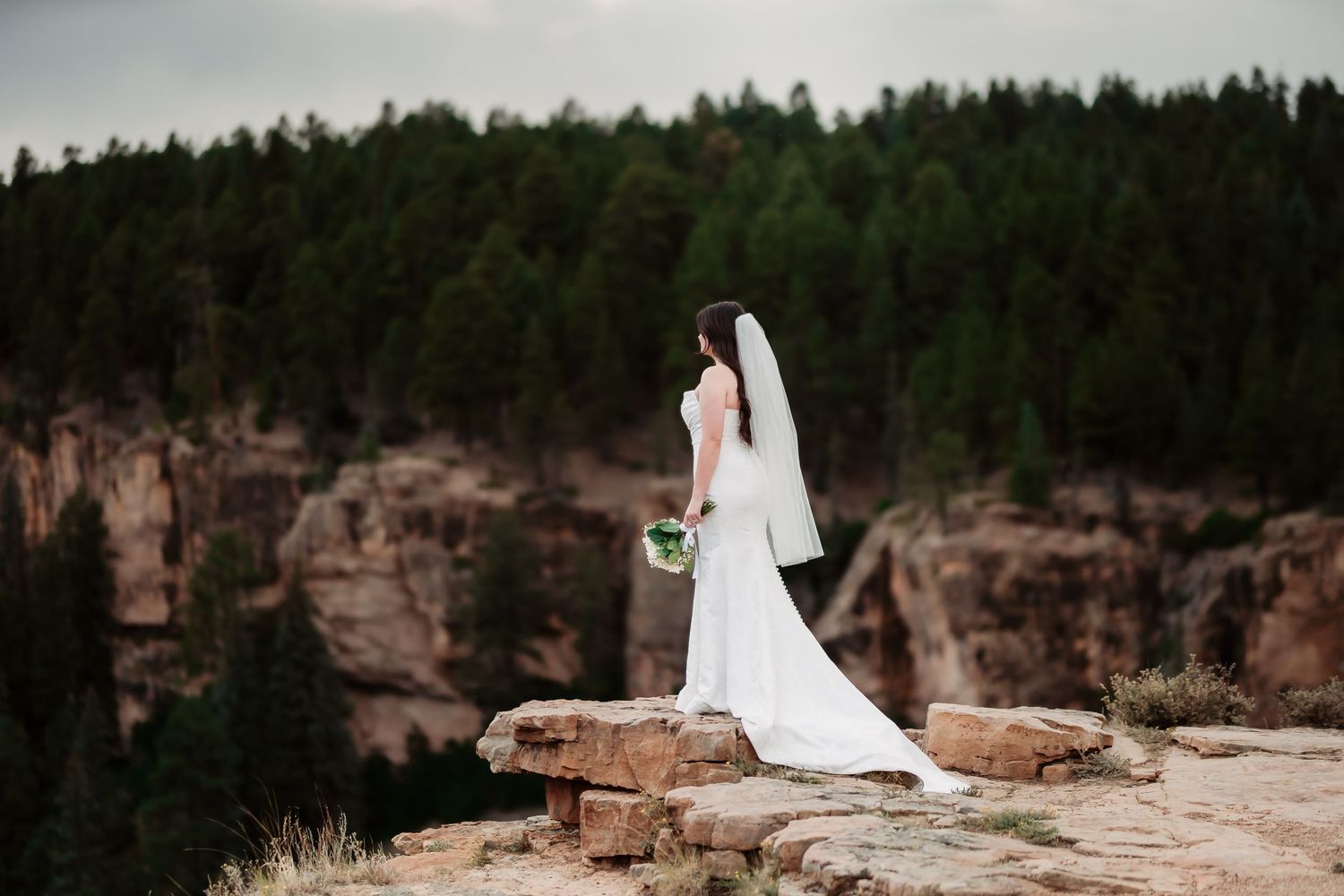 A wedding couple stands atop red rock cliffs in a dramatic outdoor mountain landscape at sunset.