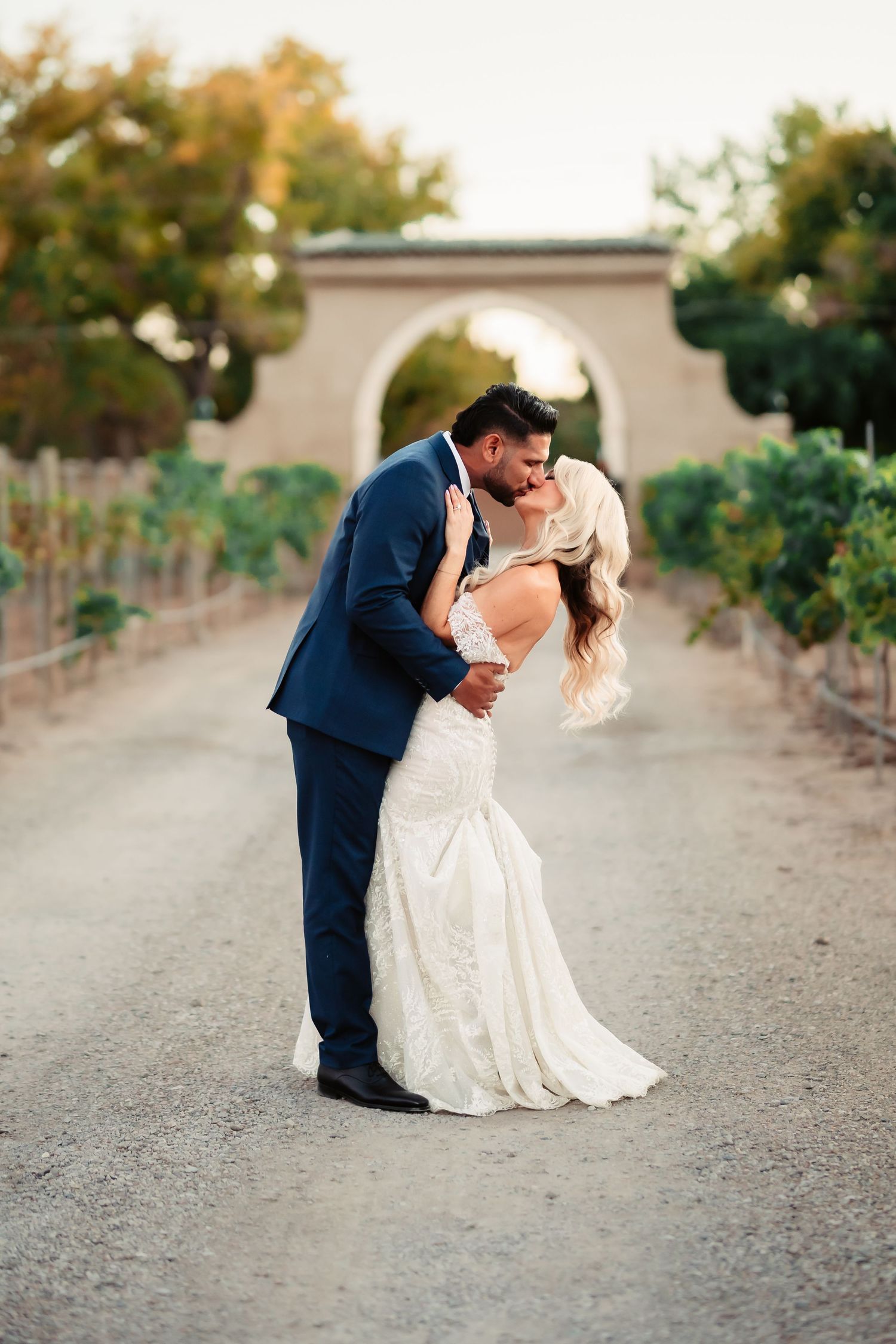 A romantic dip kiss between newlyweds on a dusty vineyard path at sunset.