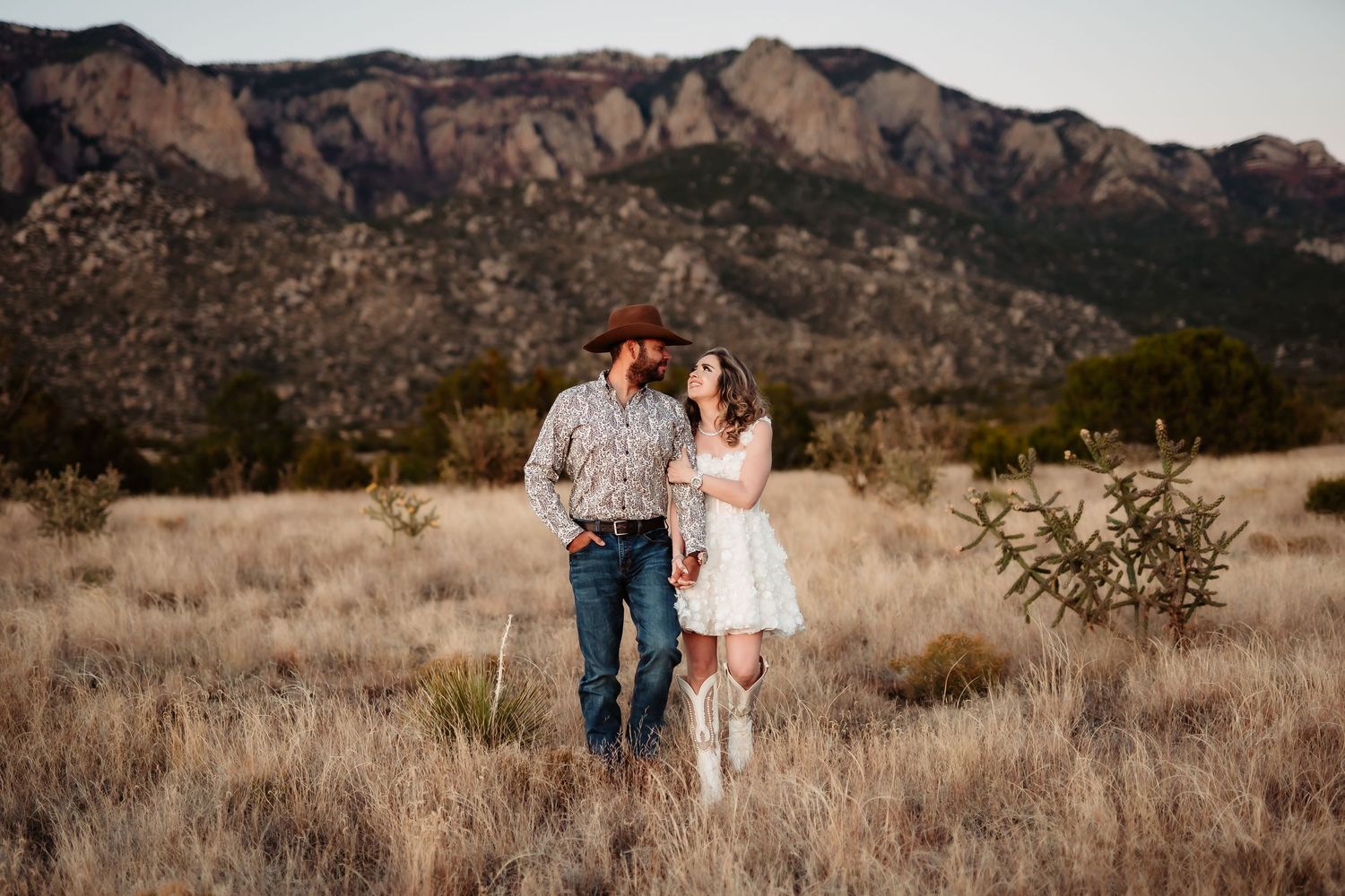 Romantic couple embraces in golden field with rocky mountain backdrop during sunset.