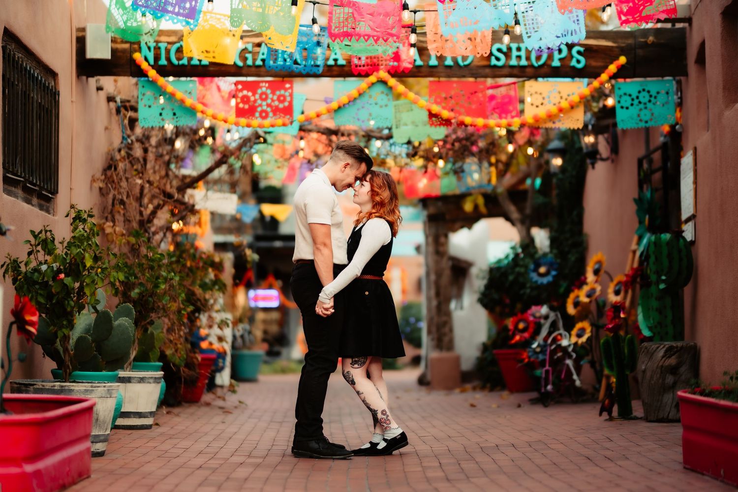 Colorful papel picado banners hang over a festive Mexican street market with vendors and shoppers.