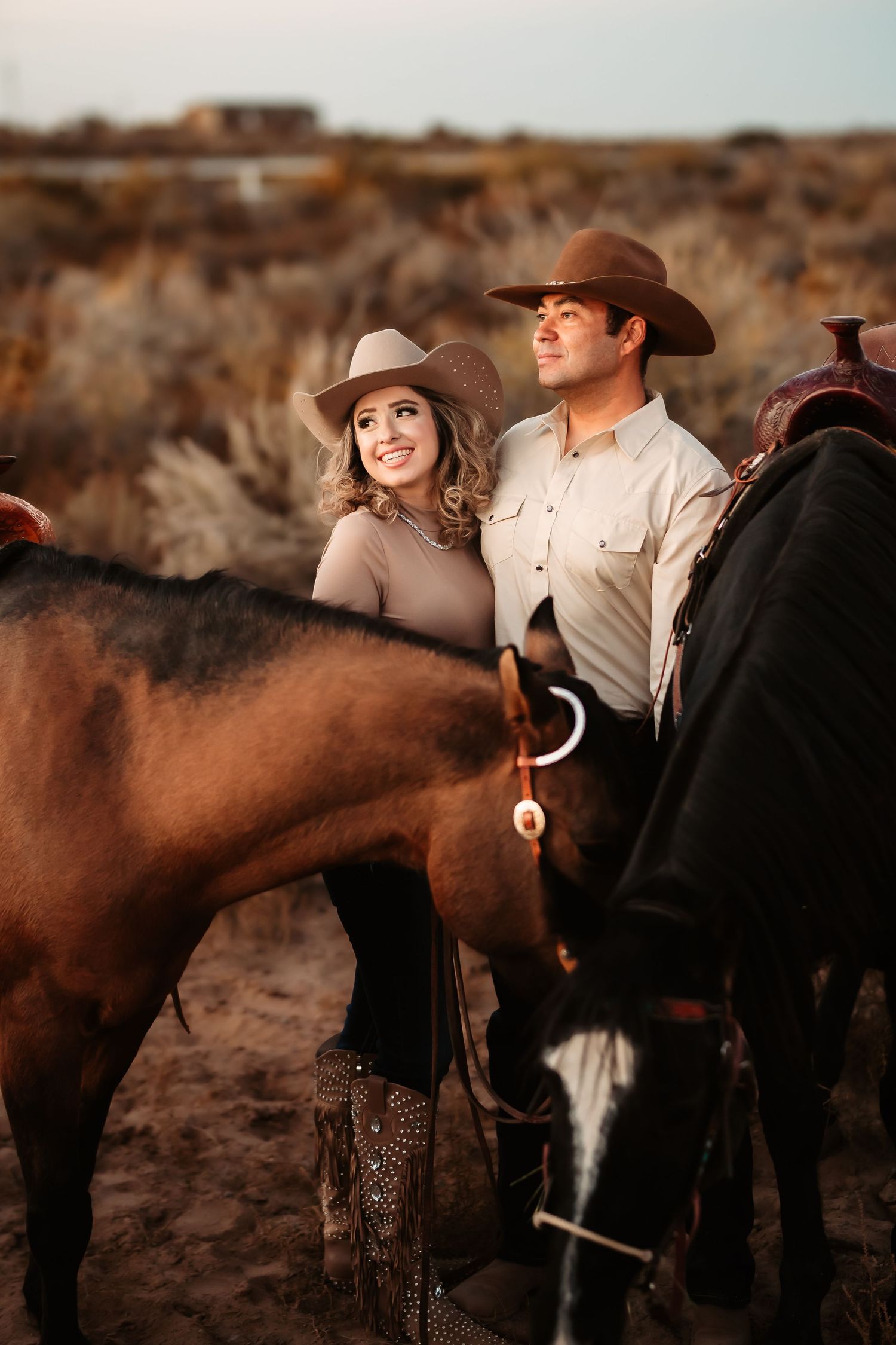 A couple in western attire stands beside their horse during a countryside photoshoot.