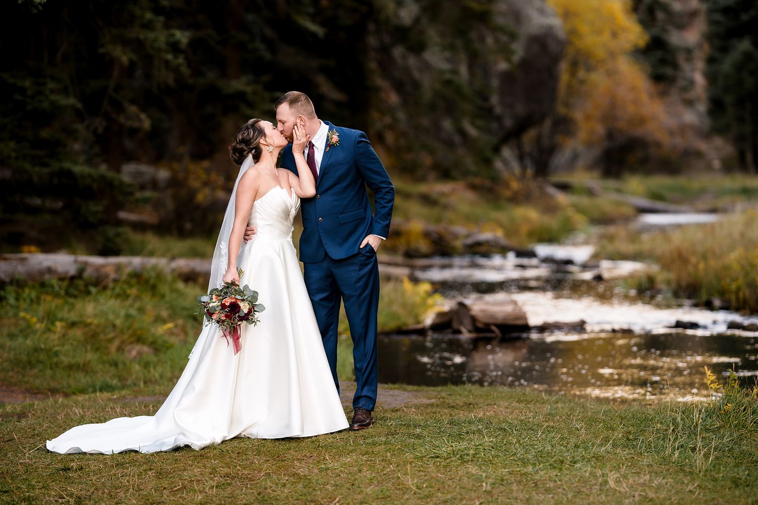 Bride and groom share a kiss outdoors near a mountain stream, surrounded by autumn foliage.
