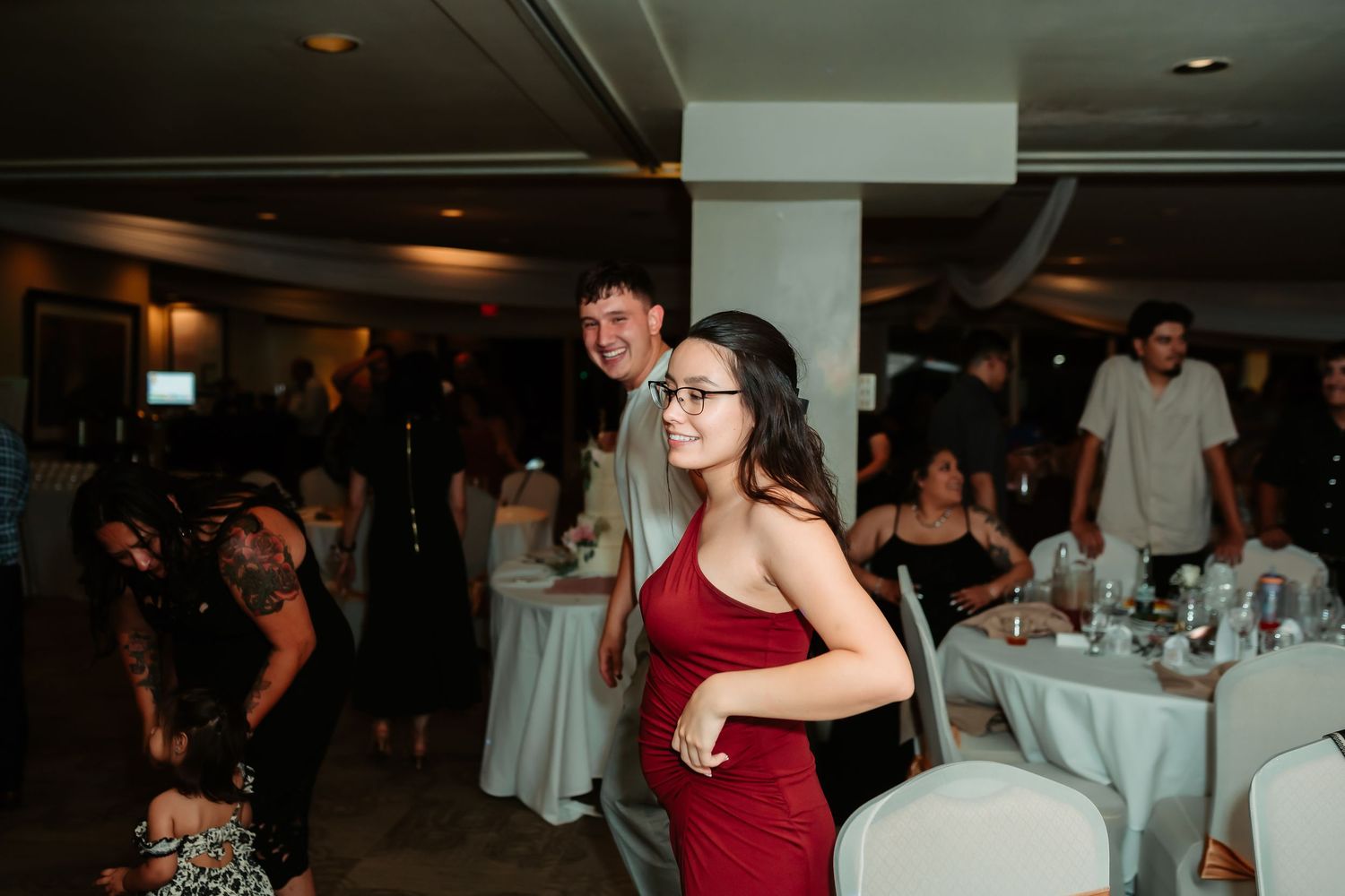 Wedding reception guests dancing and mingling among white-clothed tables in a dimly lit venue.