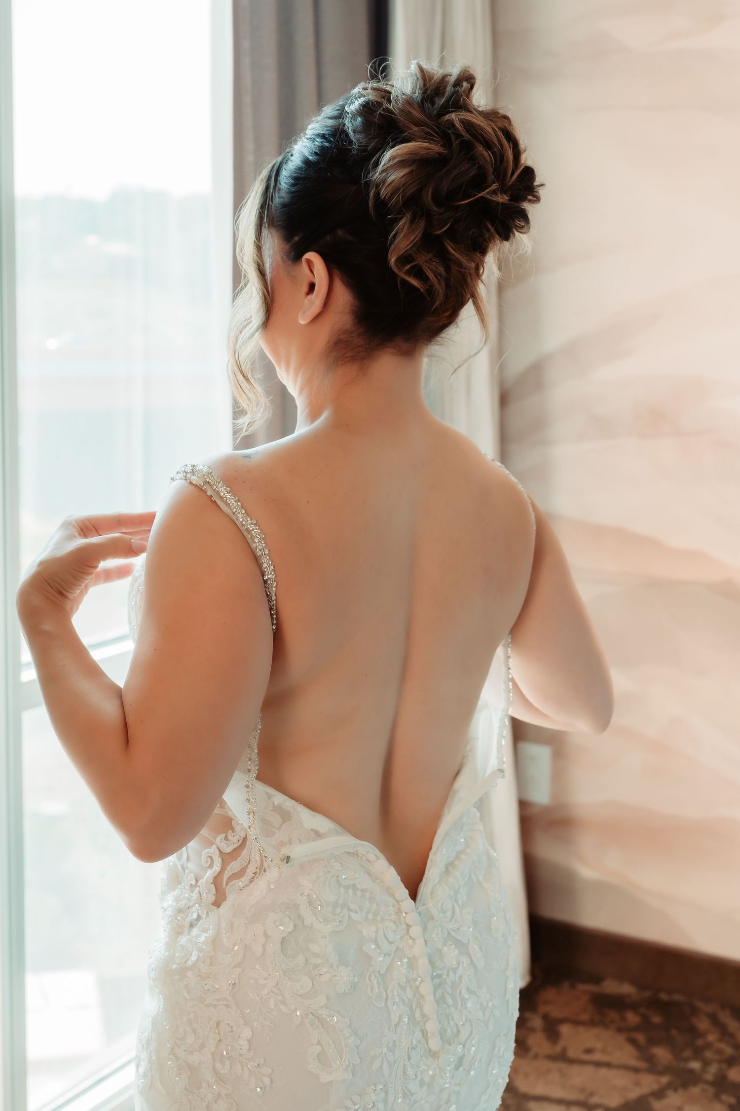 A bride in a white lace gown stands by a window as she prepares for her wedding day.