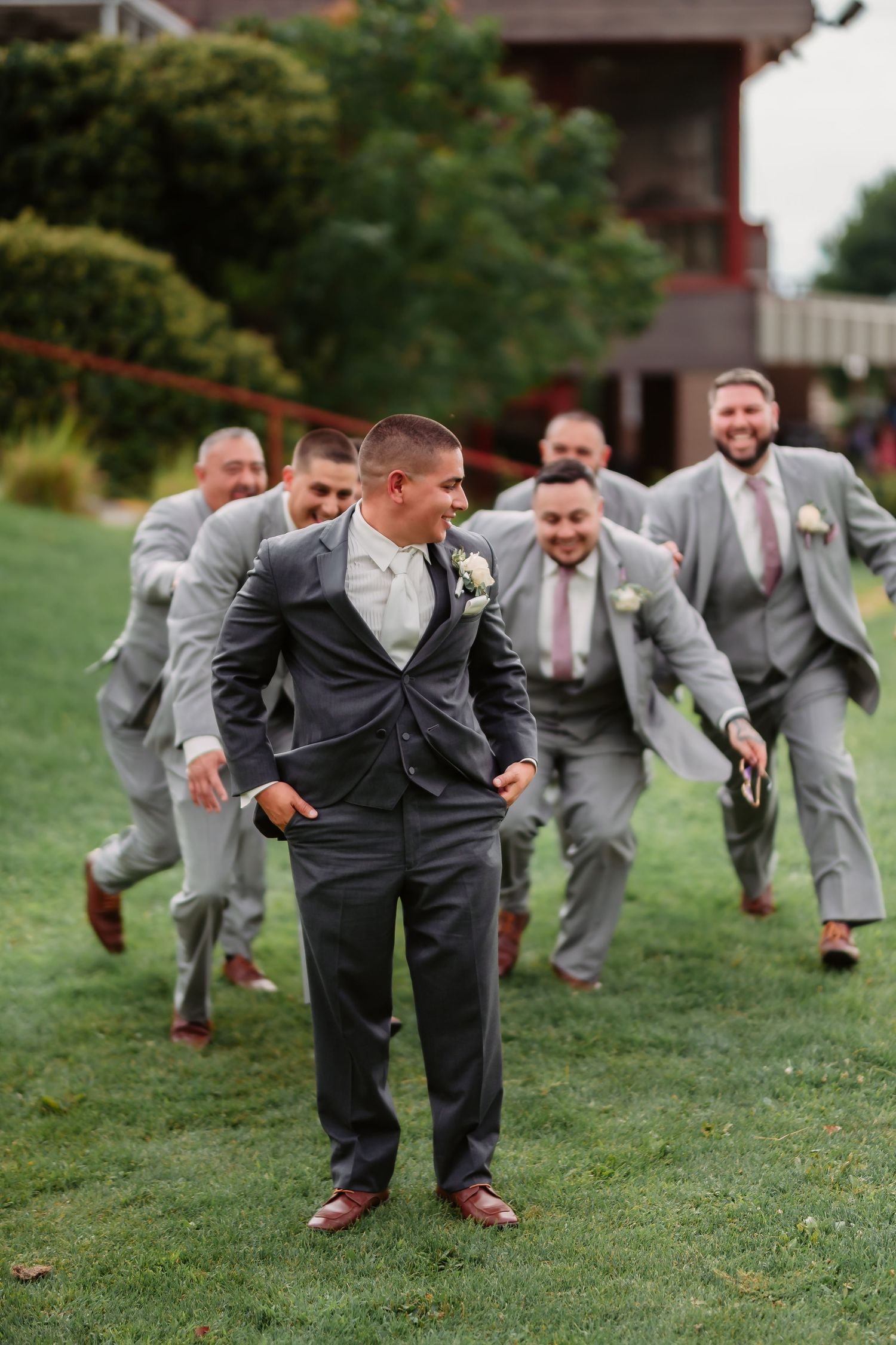 A group of groomsmen in gray suits run playfully across a green lawn behind someone in a dark suit.
