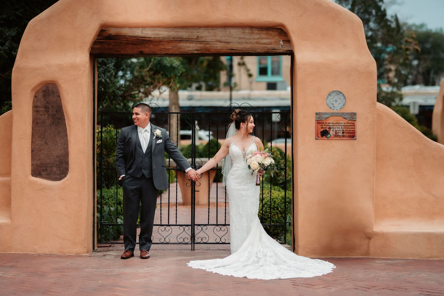 Wedding couple standing in front of terracotta colored wall and iron gates at outdoor venue.