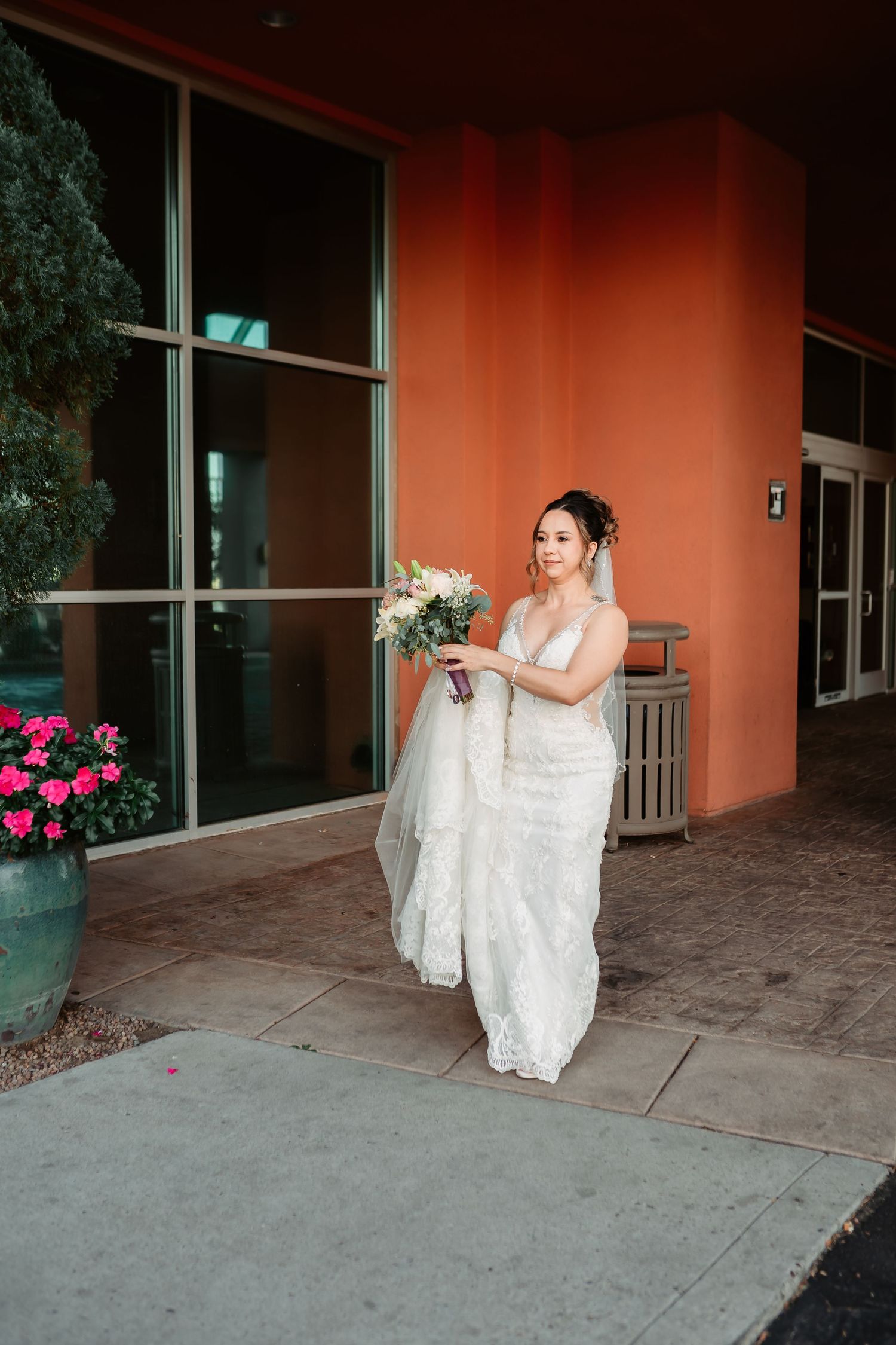 A bride in a white lace gown holds her bouquet while standing outside a building with orange walls and glass windows.