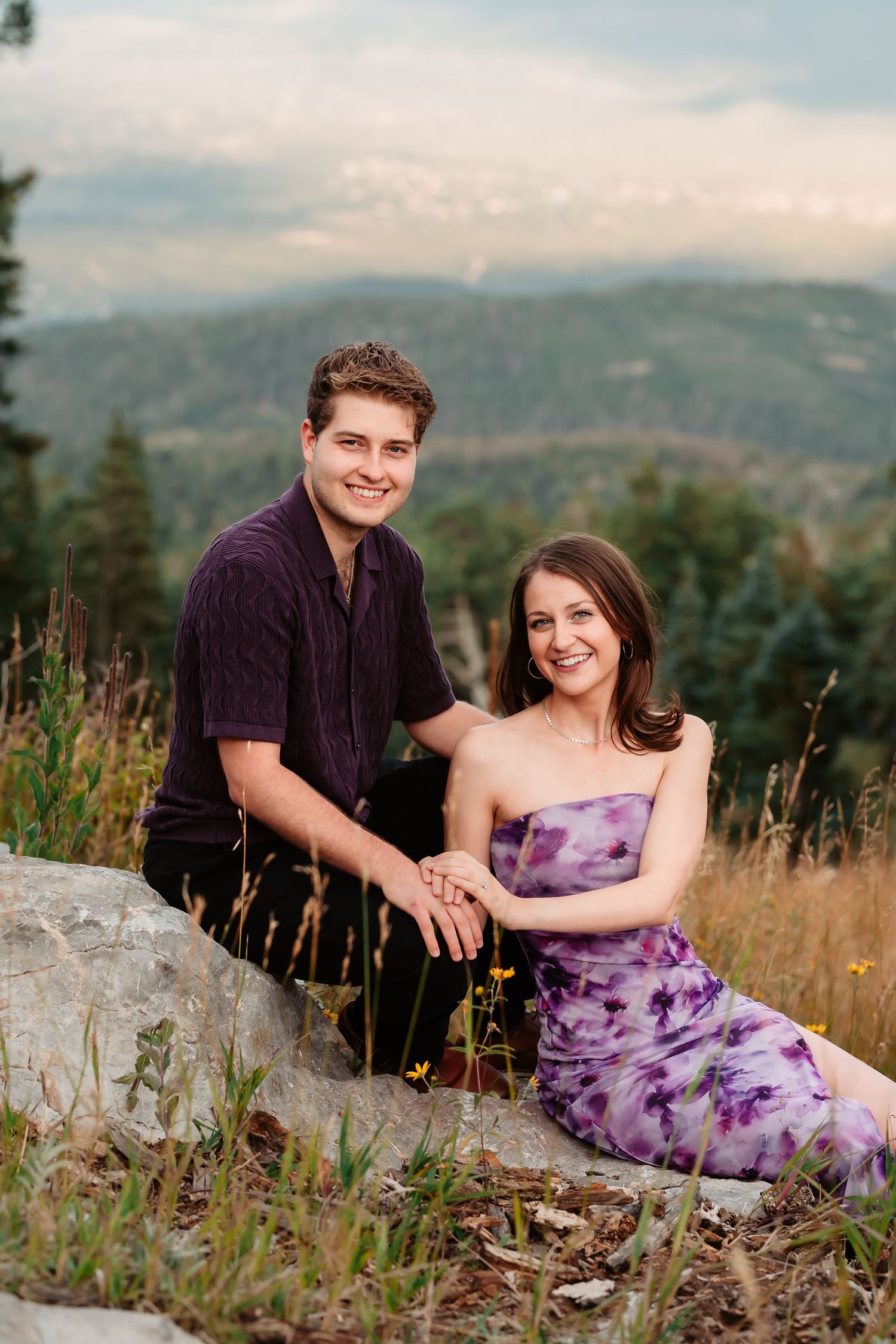 Engagement portrait in a mountain meadow with wildflowers and forest backdrop.