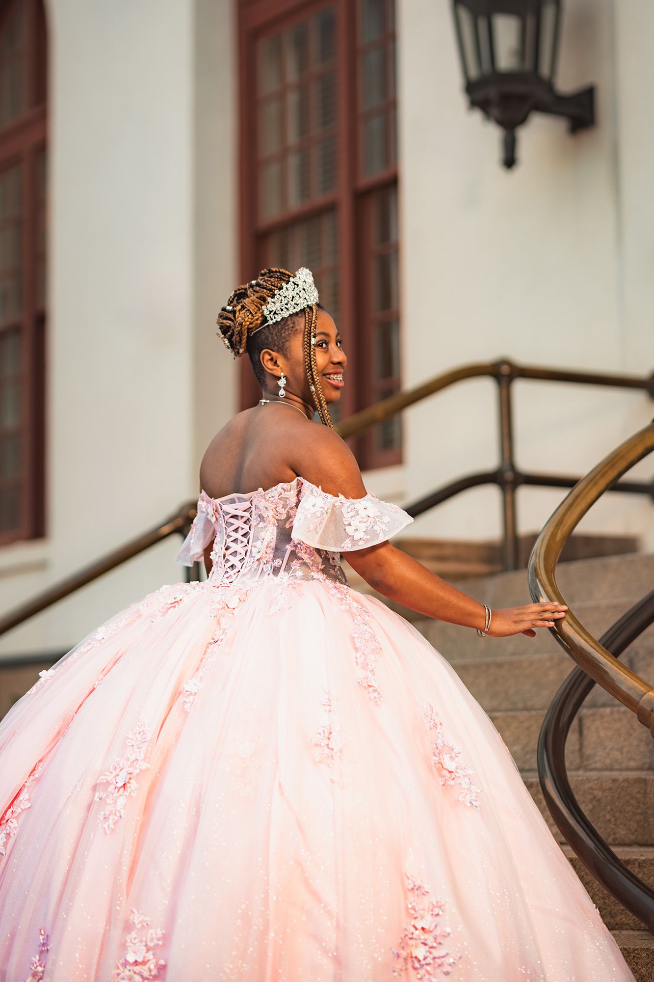 A person in a flowing pink ball gown poses gracefully on outdoor stone steps with red-trimmed windows in the background.