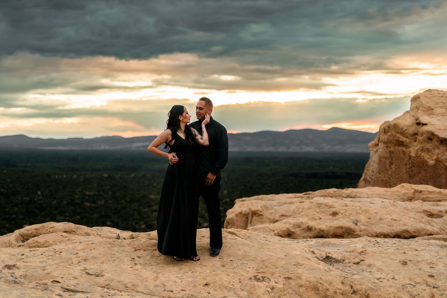 Couple in black formal attire poses on rocky desert cliff at sunset with dramatic cloudy sky and valley views.