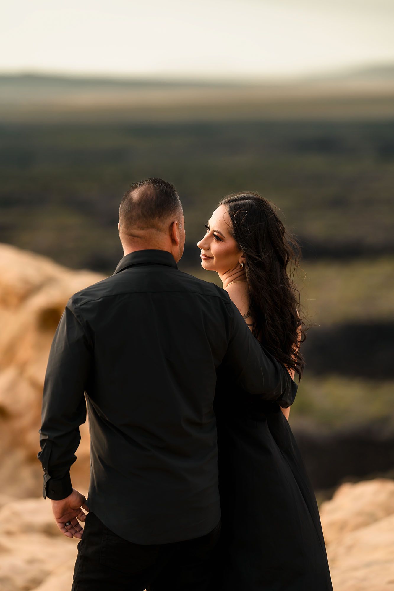 Couple in matching black outfits standing on rocky overlook, woman smiling back at camera with scenic landscape behind.