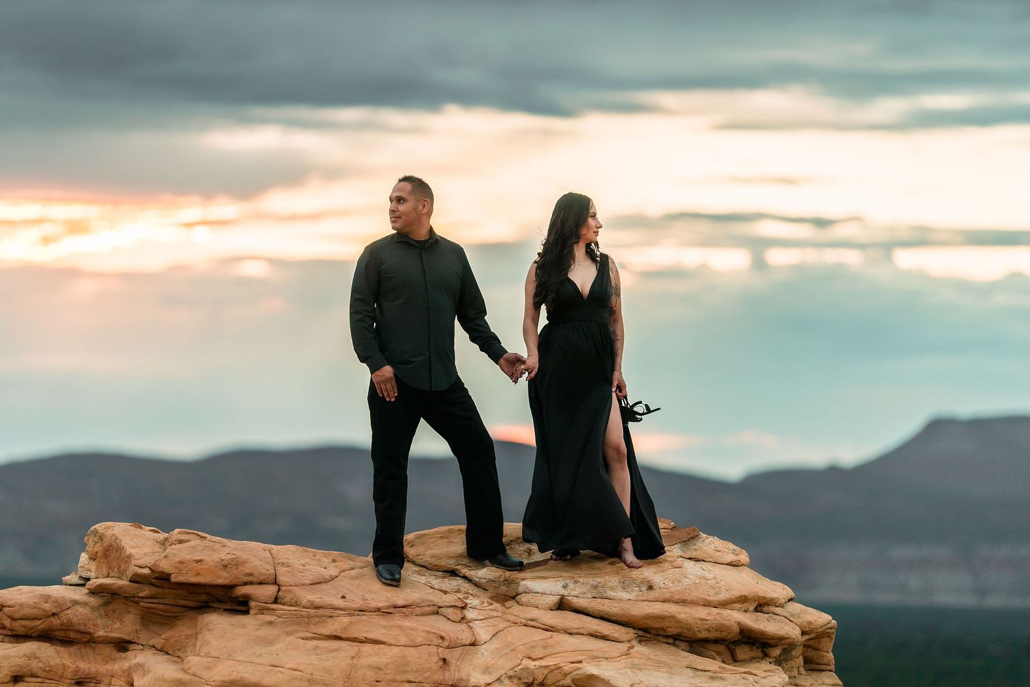 Couple in elegant black outfits holding hands on desert rock formation at sunset.