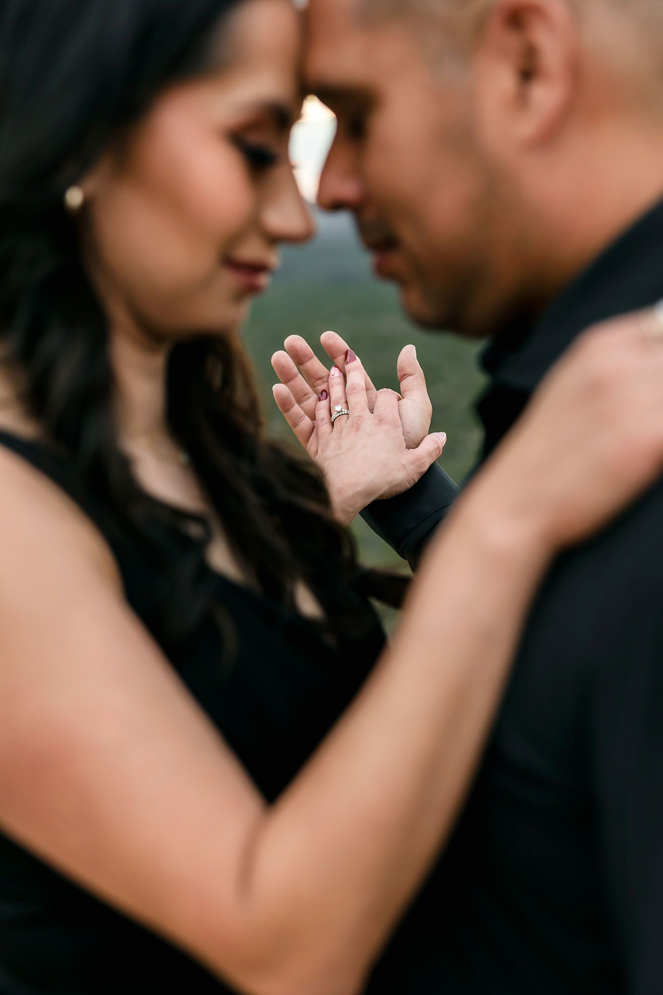 Engaged couple in black touching foreheads while woman displays ring in outstretched hand.