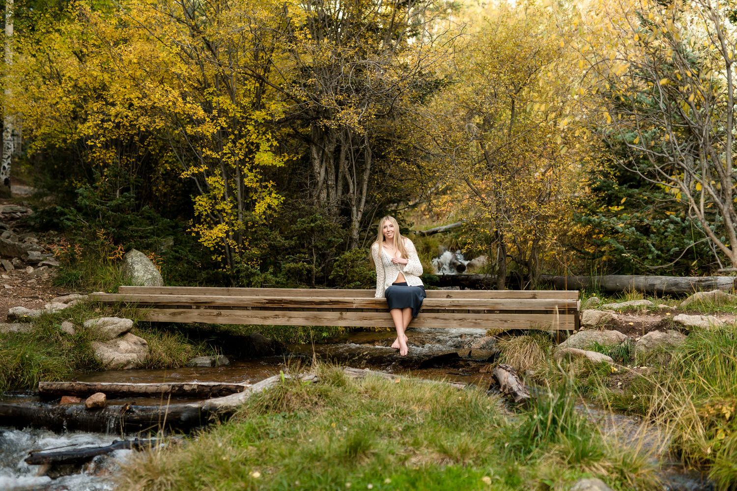 Blonde woman sitting on a wooden bridge over a stream surrounded by autumn foliage in a forest.