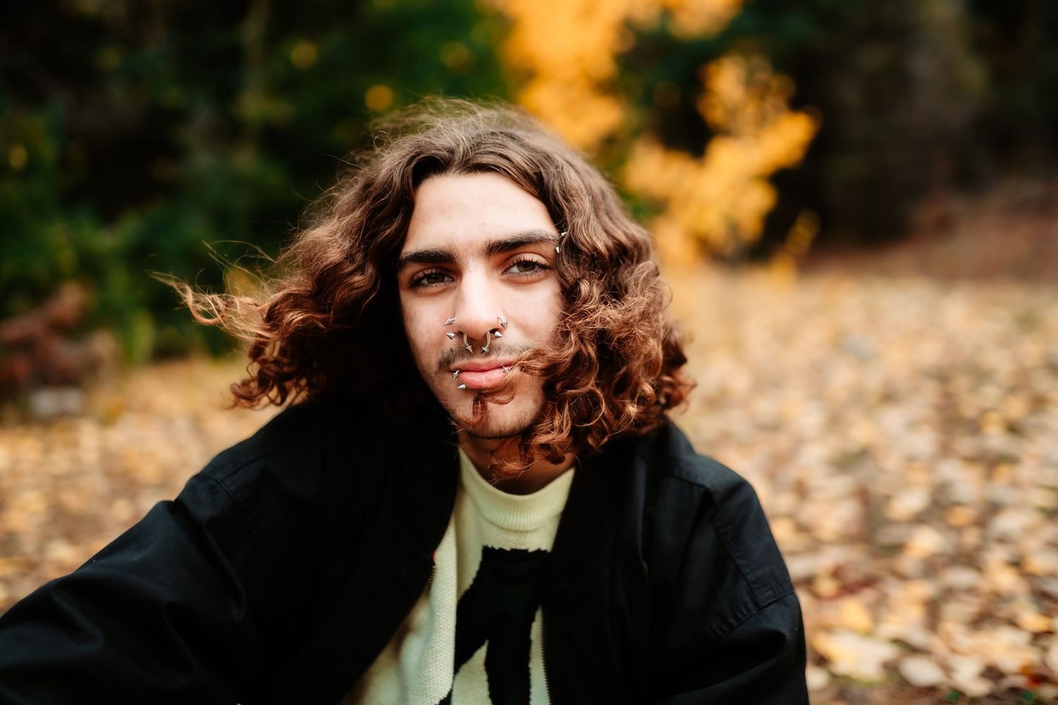 A person with long curly hair sits outdoors among autumn leaves during golden hour.
