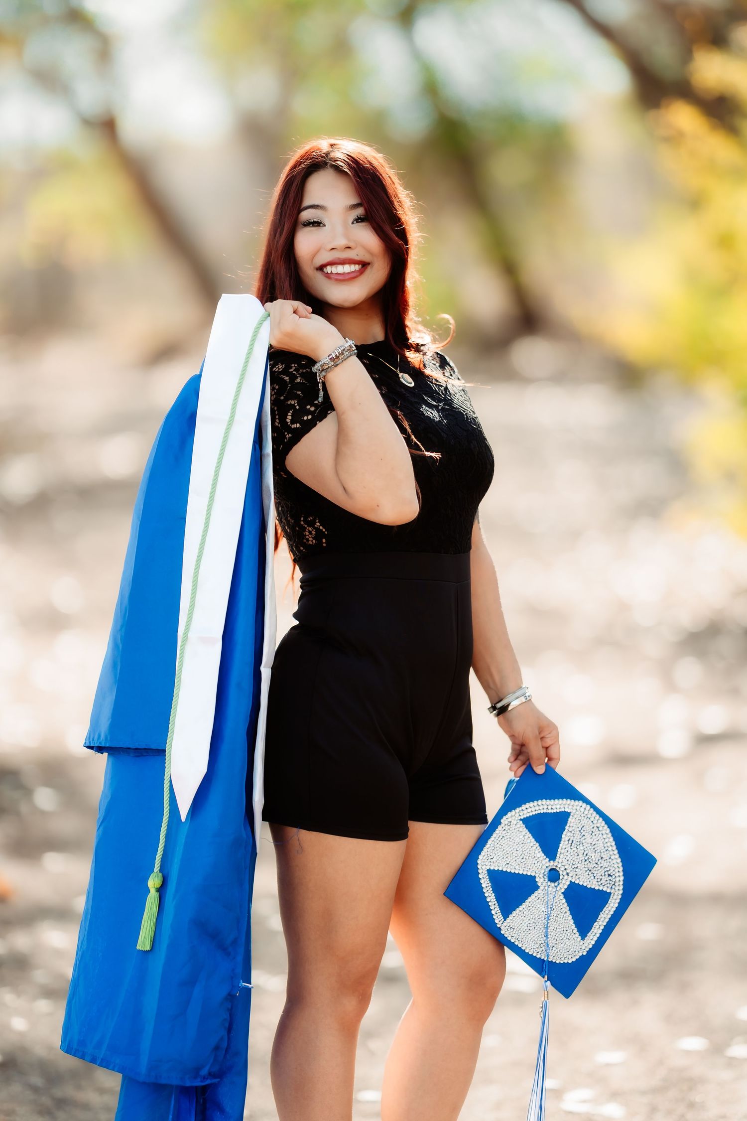 Graduate in black dress holding blue graduation gown and diploma standing outdoors in natural setting.