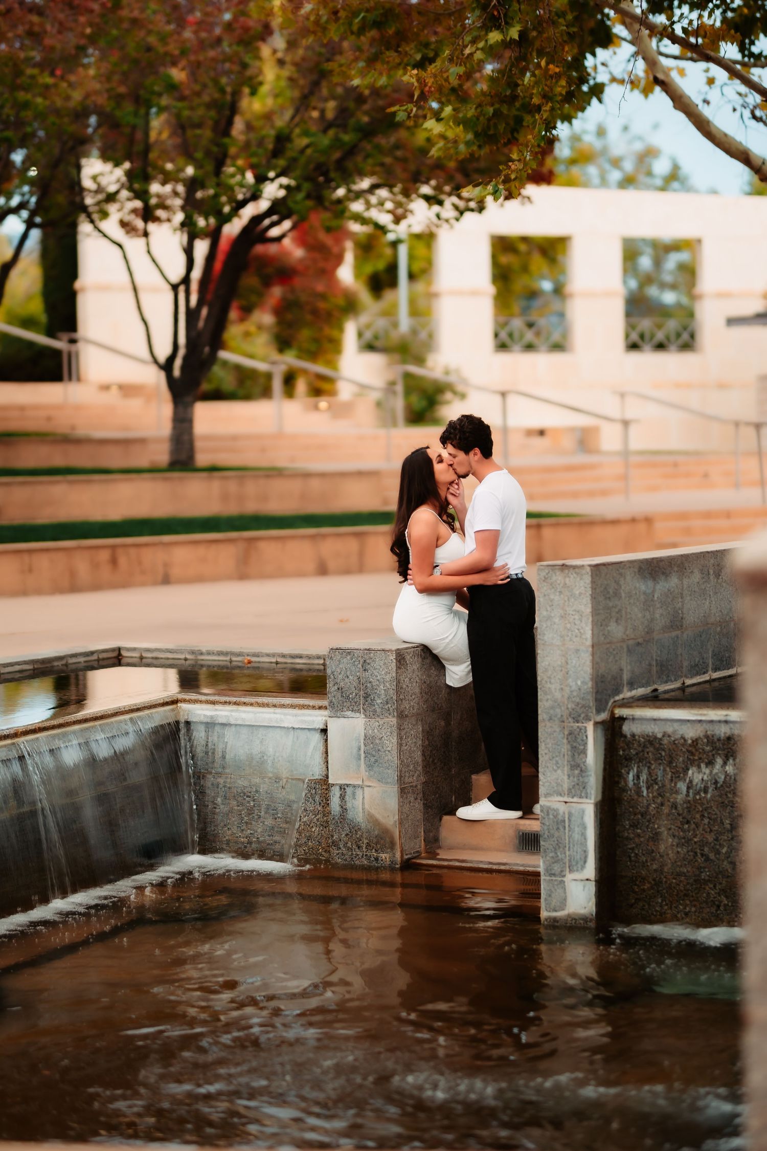 A couple sits intimately by a reflecting pool surrounded by autumn trees and stone architecture at sunset.