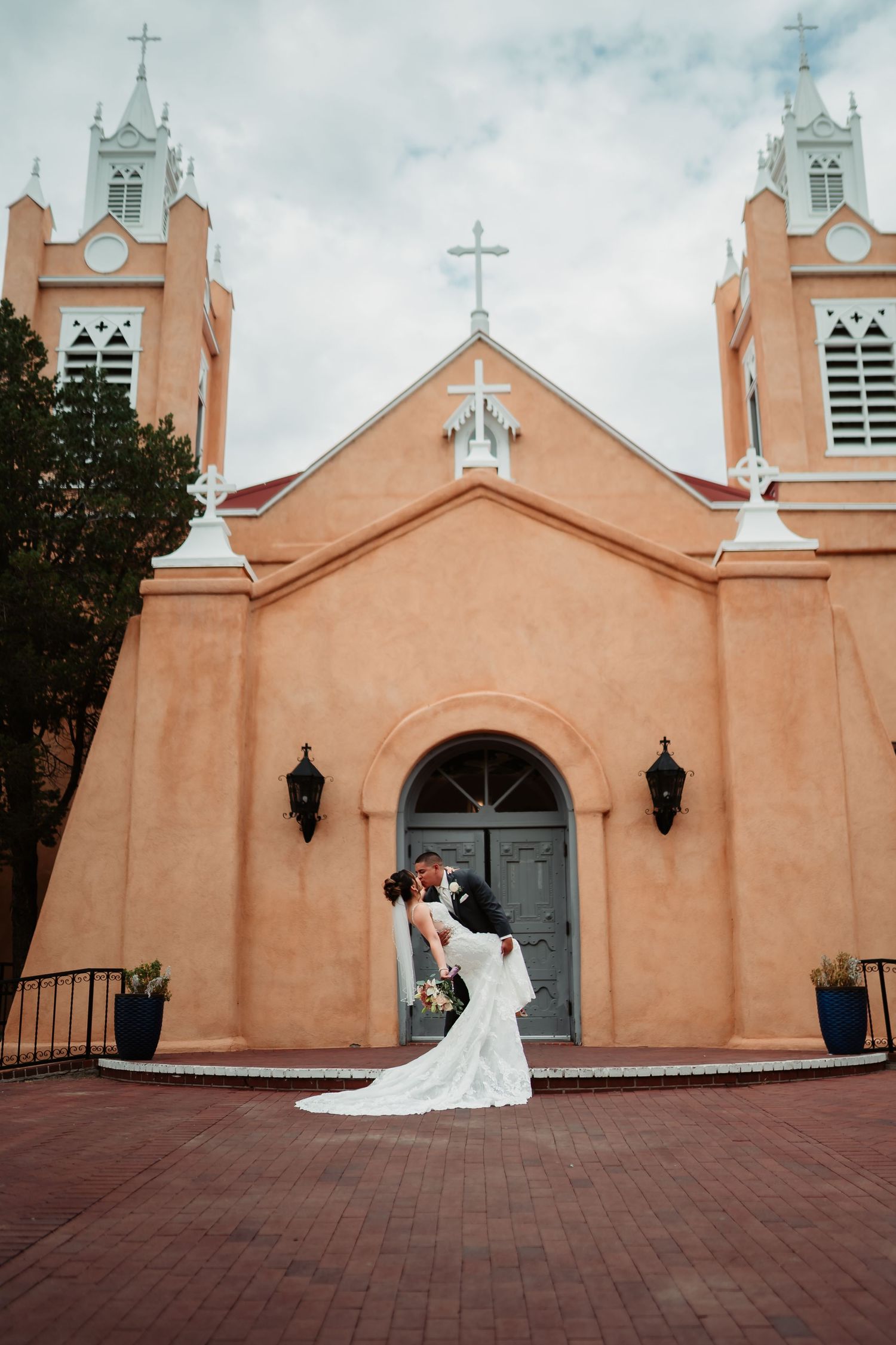 A couple poses in front of a peach-colored Spanish colonial church with white crosses and bell towers in New Mexico.