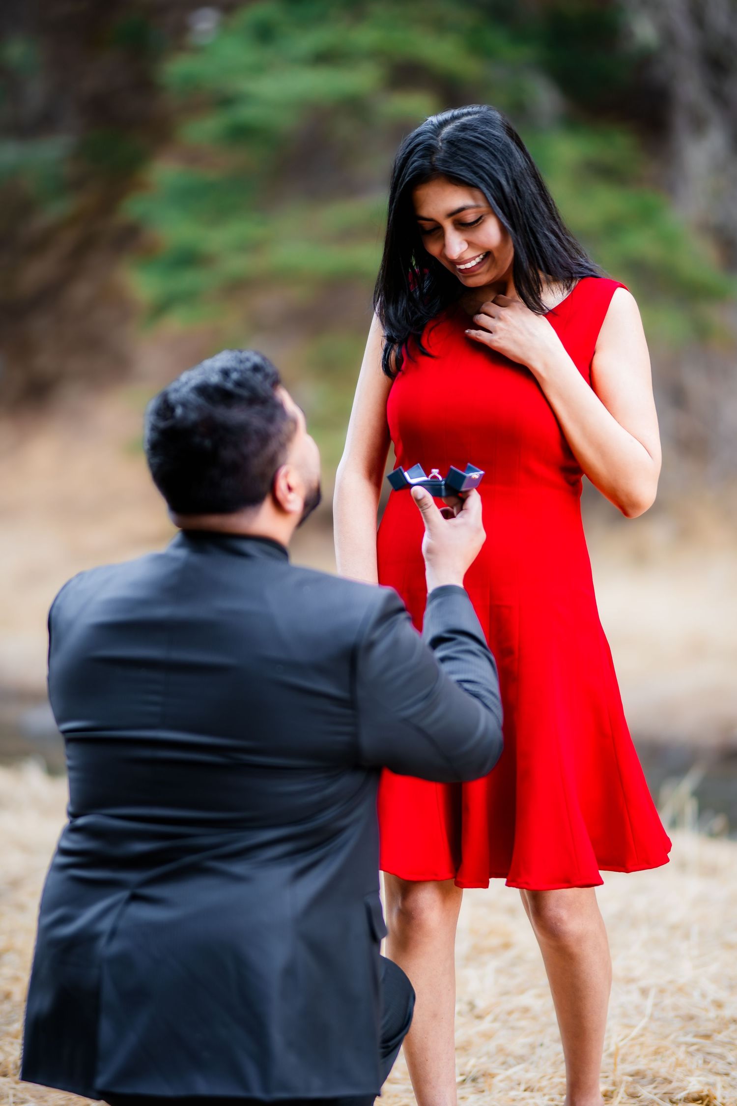 A couple shares a romantic proposal moment outdoors with a red dress and black suit.
