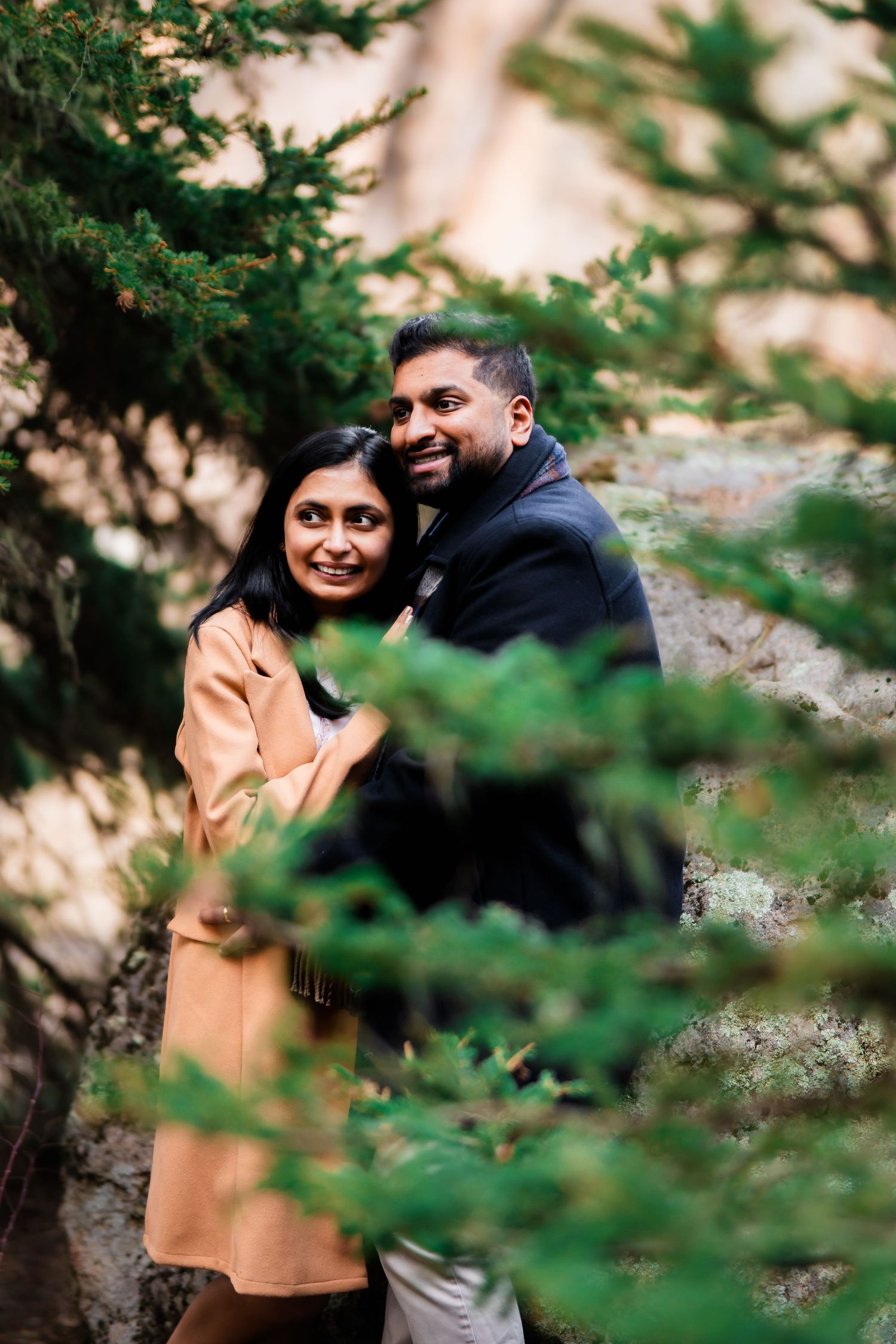 A joyful couple posing together between pine branches during their outdoor photoshoot.