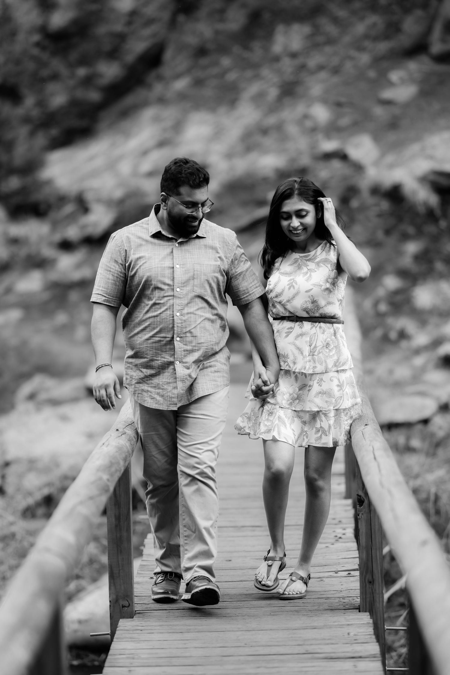 A couple walks hand in hand across a wooden footbridge in a black and white nature setting.