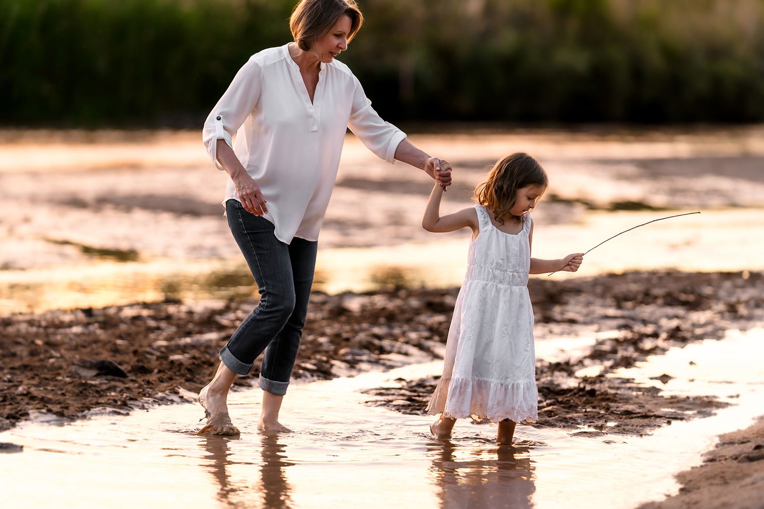 Woman and girl wading in shallow water at sunset, holding hands while child holds a fishing rod.