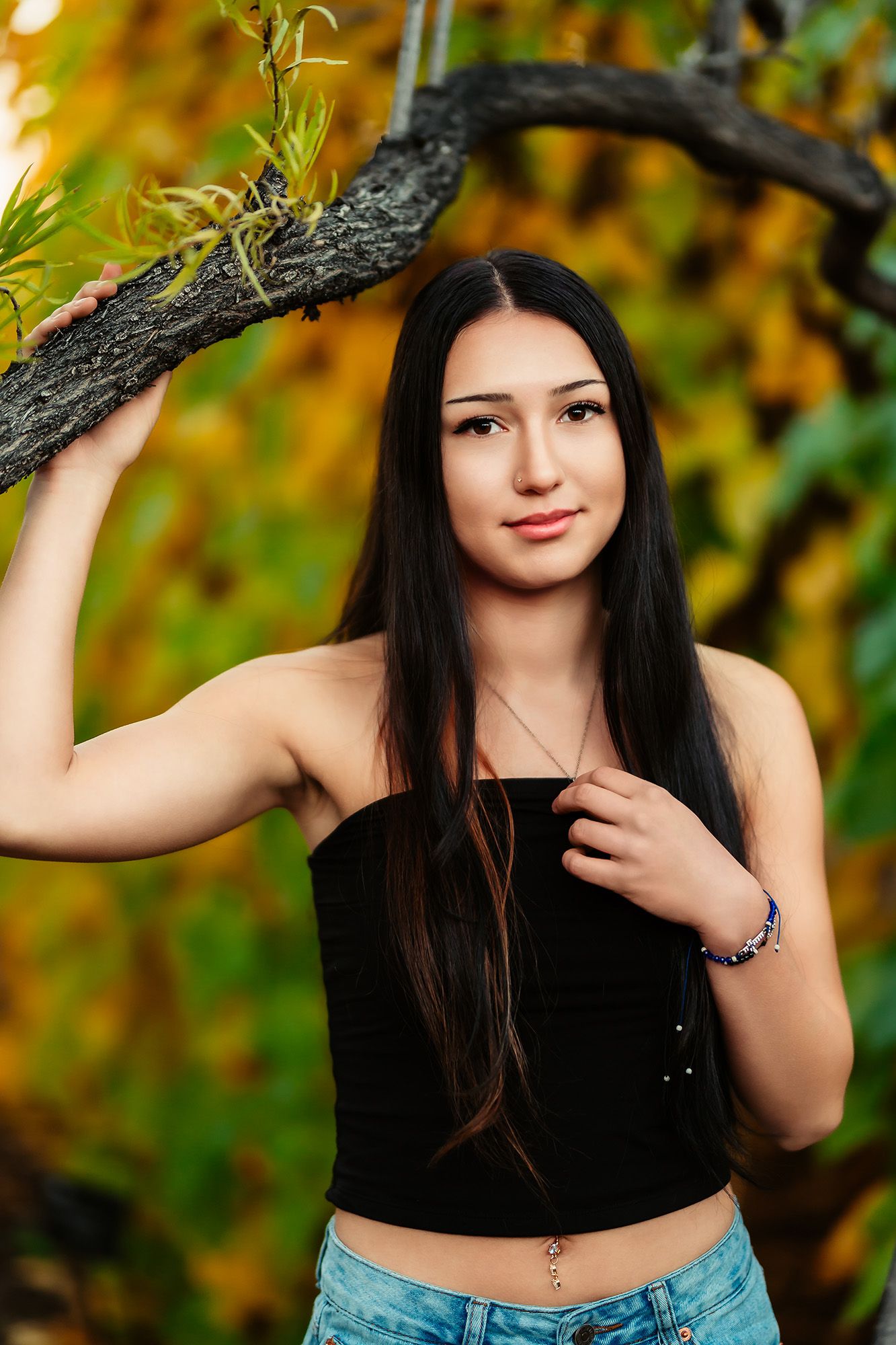 A person poses in a black top against an autumn backdrop with golden foliage and tree branches.