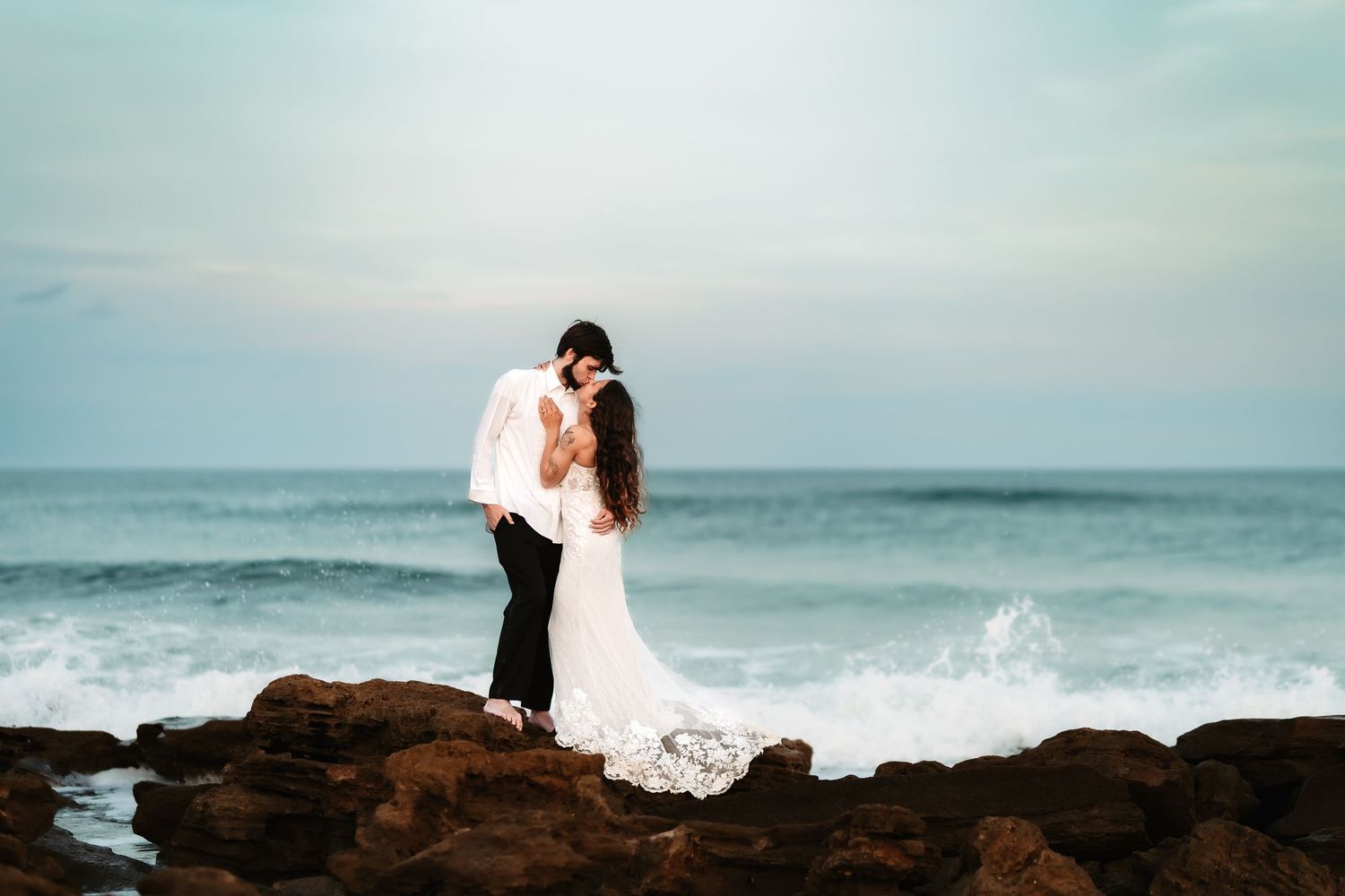 A couple stands on rocky cliffs overlooking the ocean as waves crash against the shore during a romantic photoshoot.