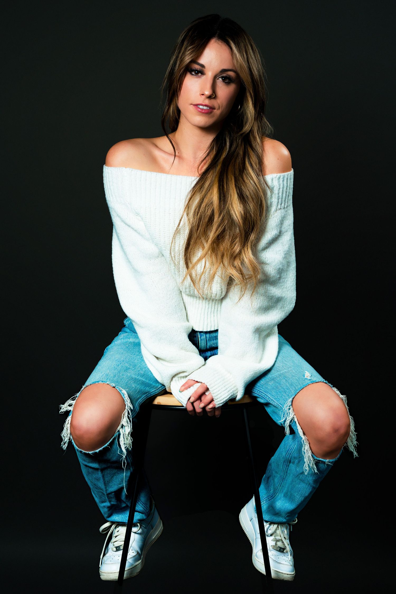 Fashion portrait with off-shoulder white sweater, distressed blue jeans and sneakers against dark background.