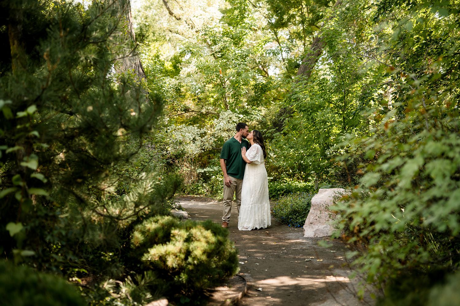 Couple sharing a romantic embrace on a garden path surrounded by lush green trees and foliage.