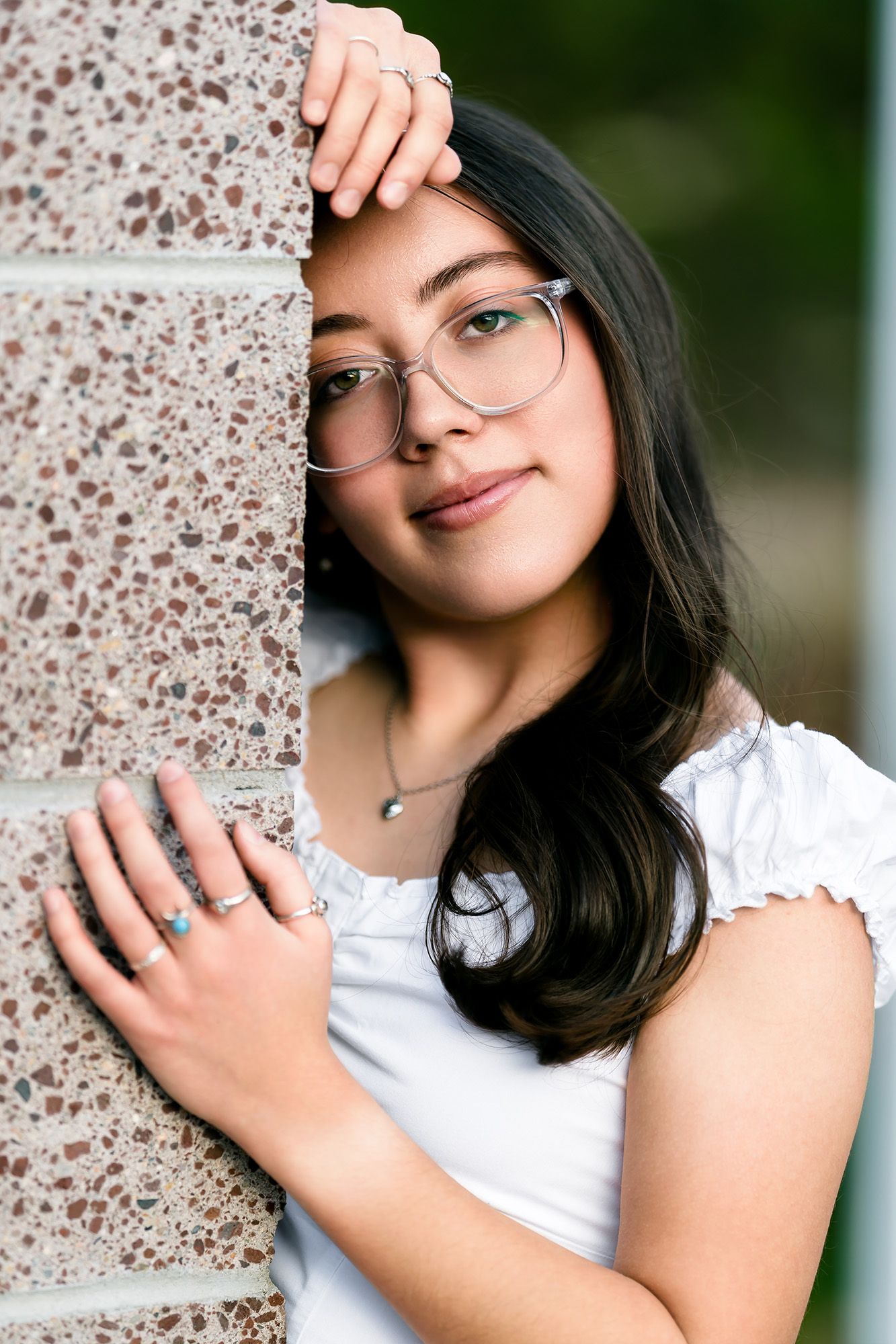 Portrait of someone in glasses and white top leaning against a textured wall with natural lighting and soft smile.