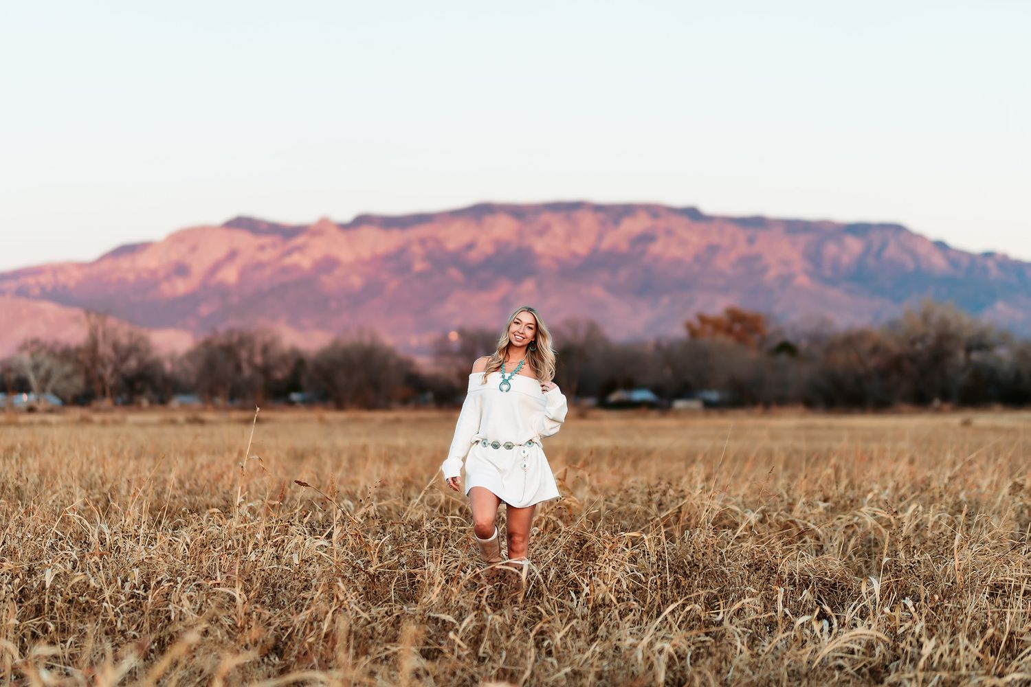 Woman in white dress and red boots standing in golden field with mountain backdrop at sunset.