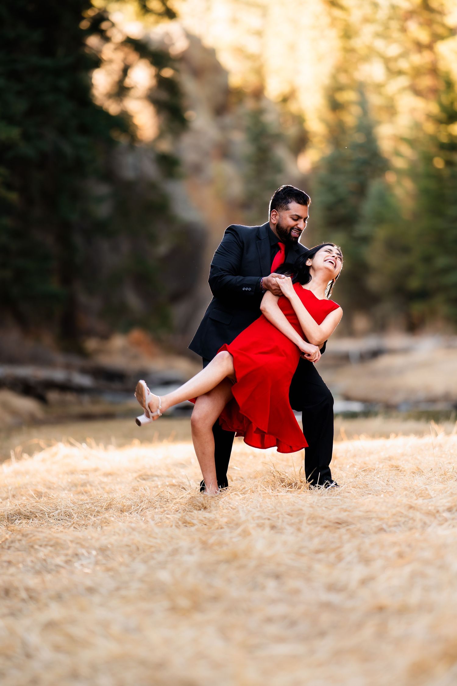 A couple in formal attire dances romantically in a sun-lit outdoor setting with evergreen trees in the background.