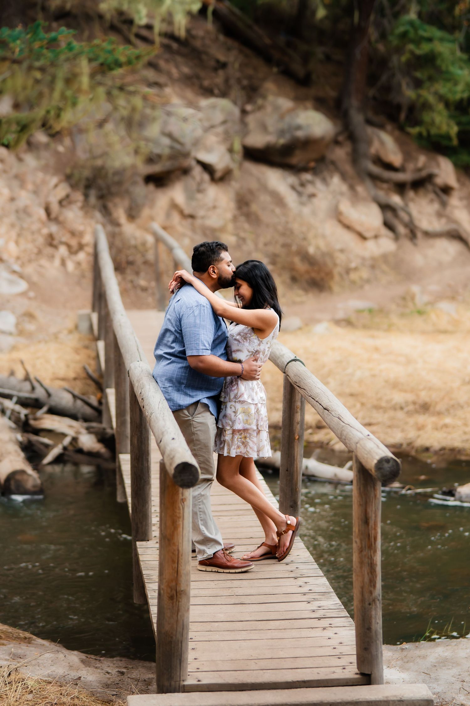 Couple embracing on rustic wooden bridge over creek with rocky cliff backdrop.