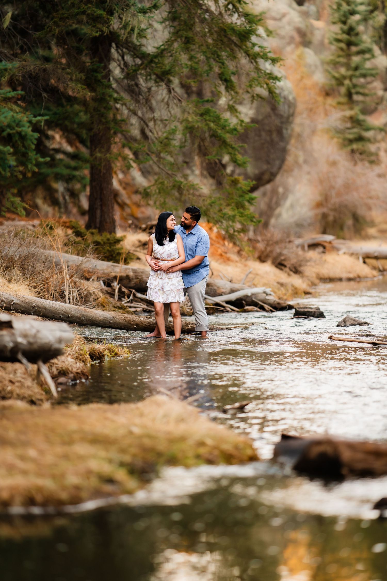 A couple stands together in shallow water surrounded by rocky terrain and evergreen trees.
