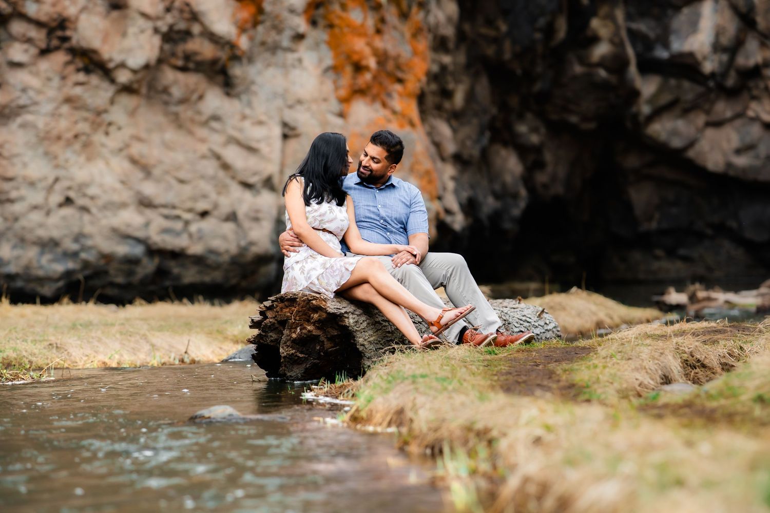 Couple sits together on rock formation by mountain stream with cliffs in background.