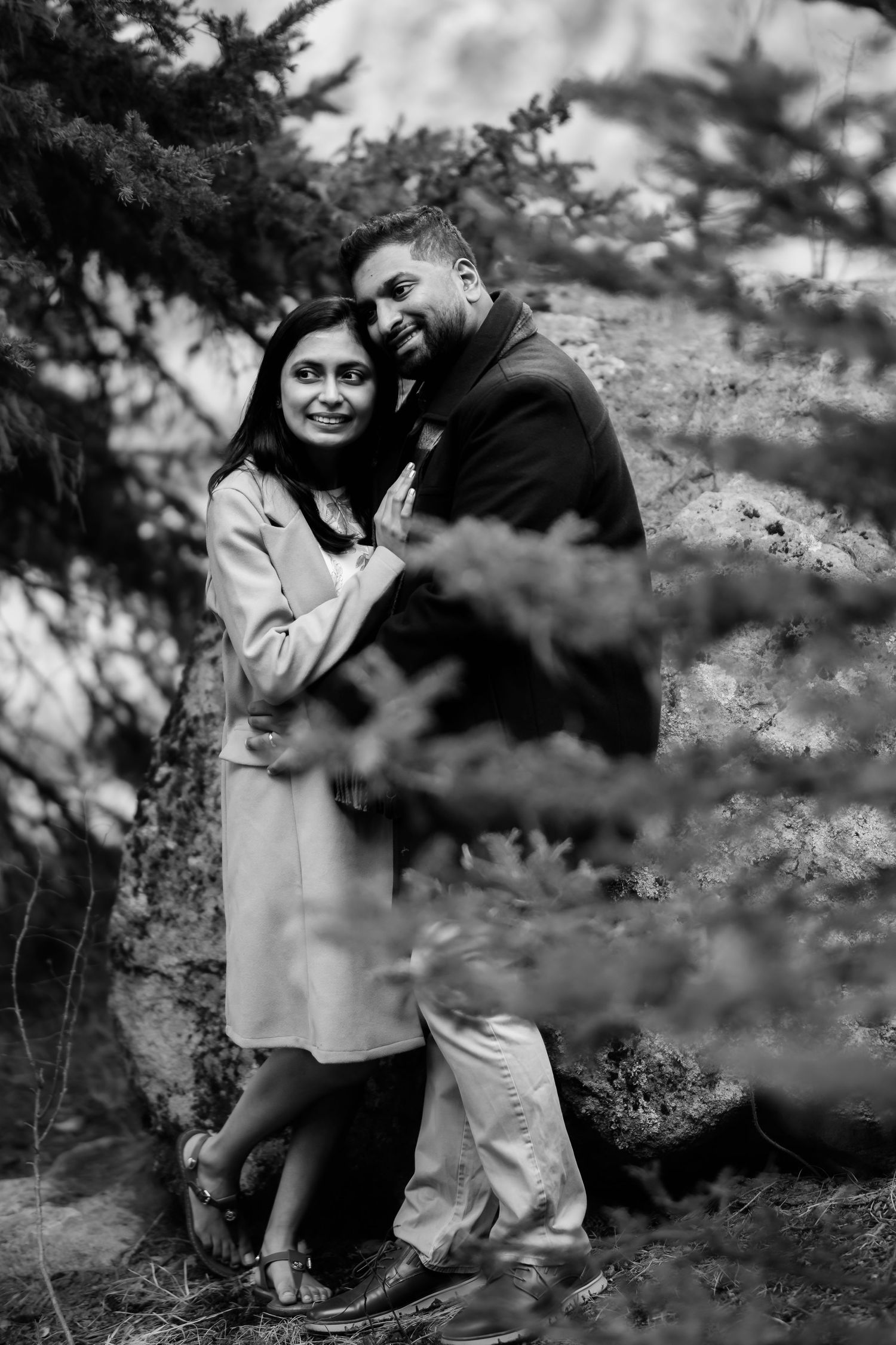 A black and white portrait of a couple embracing among evergreen trees in a natural setting.