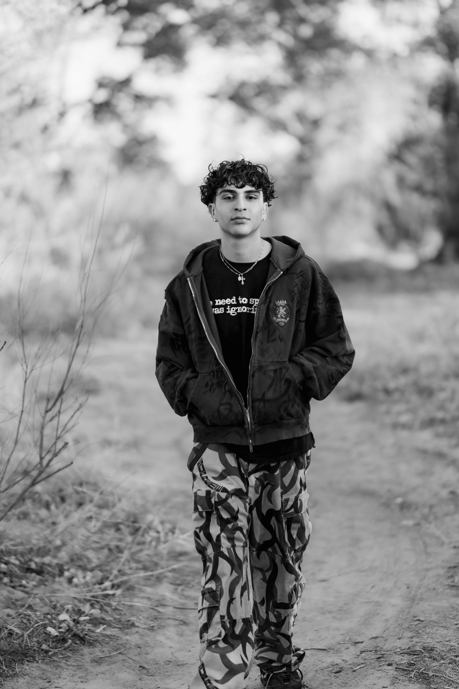 Black and white portrait of person in dark hoodie and patterned pants standing on outdoor path.