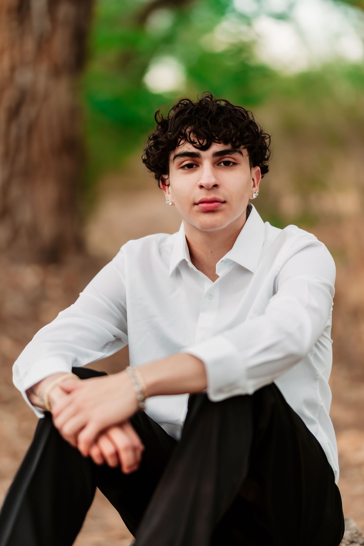 Portrait in an outdoor setting of someone in a white button-down shirt and dark pants sitting on the ground against a blurred background.
