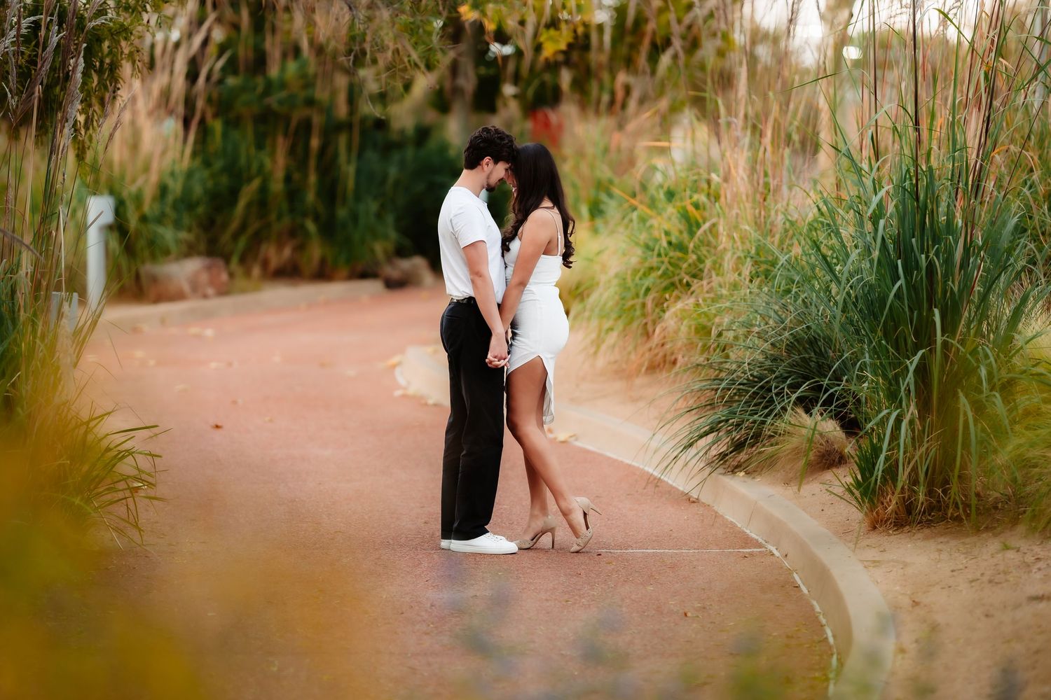 Couple embraces in a romantic moment along a winding dirt path surrounded by tall grass at sunset.