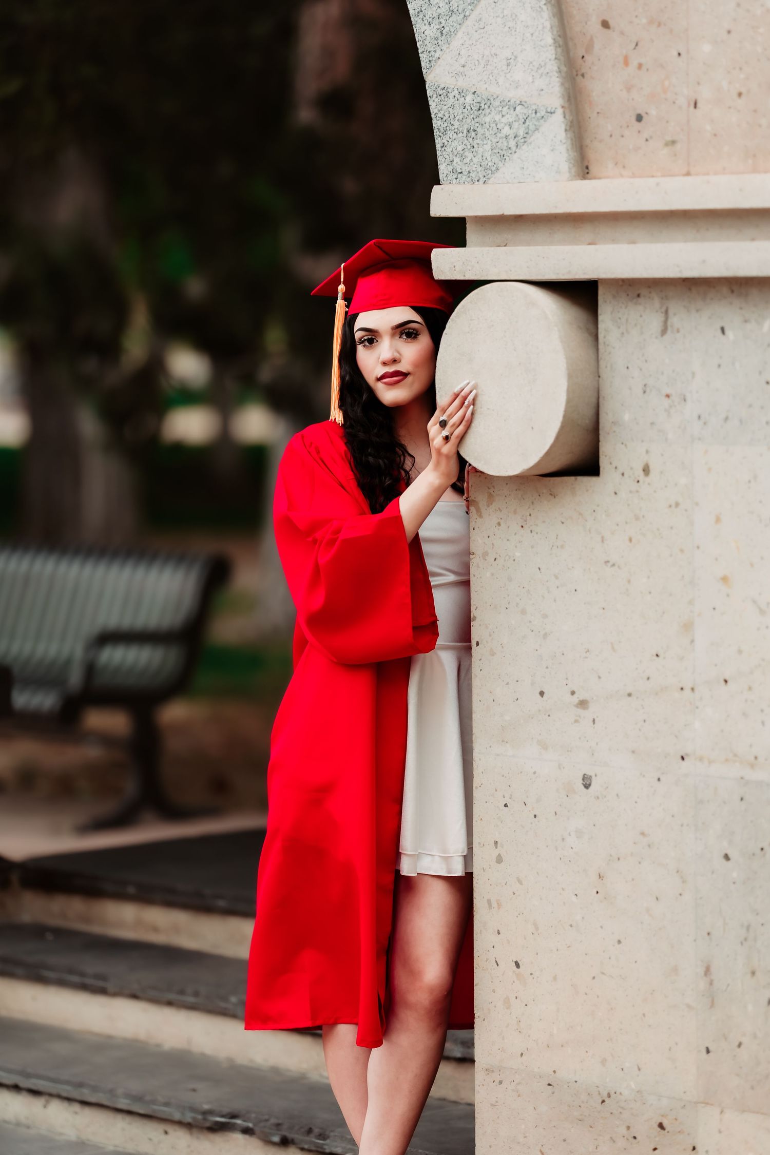 A figure in a bright red graduation gown and cap poses artistically against a white stone column outdoors.