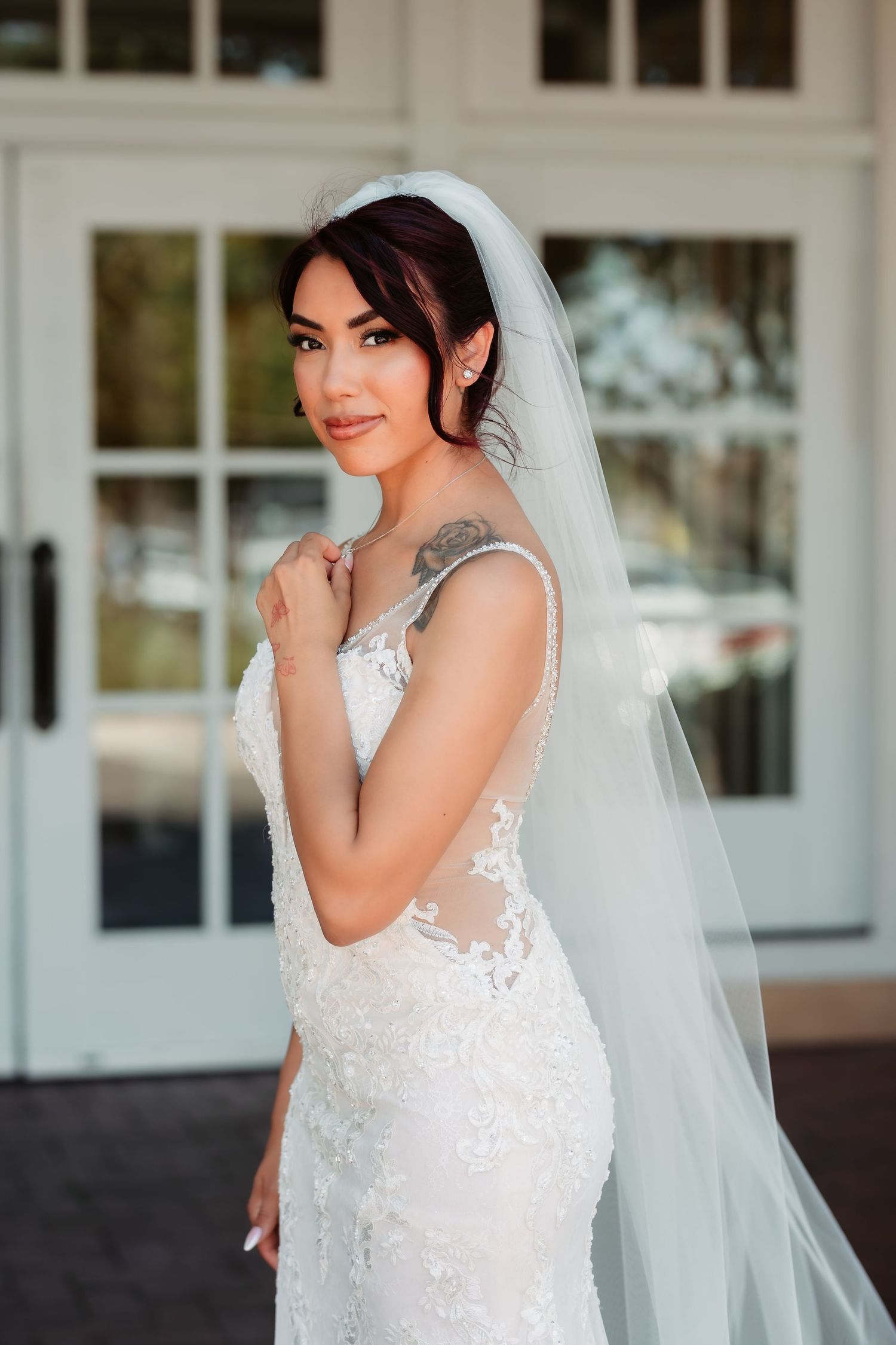 A bride in a white lace wedding dress and veil poses elegantly against a white building exterior.